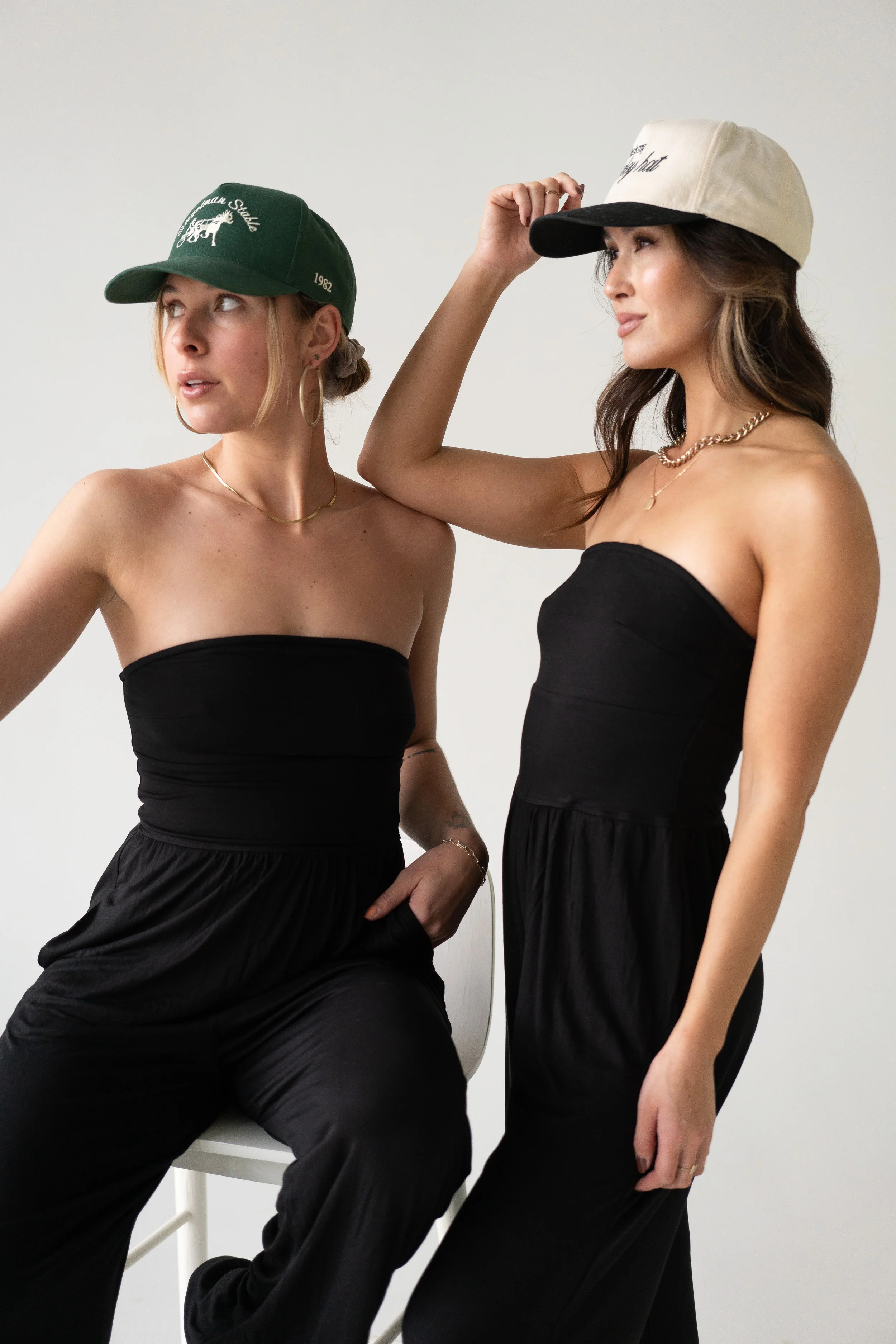 Two women in black strapless outfits and caps, posing against a plain white background.