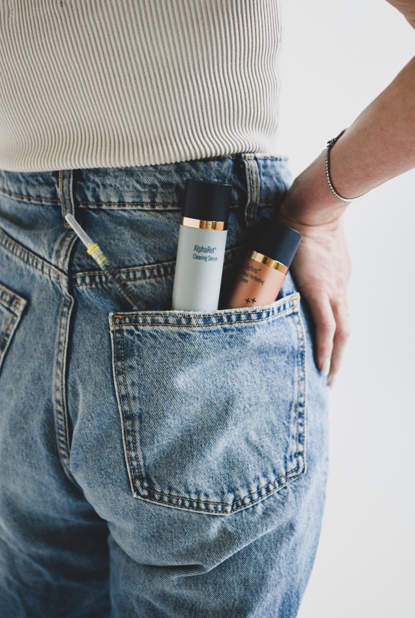Close-up of a person's back wearing blue jeans and a ribbed beige top, with skincare products in the back pocket of the jeans, including a serum and cream.