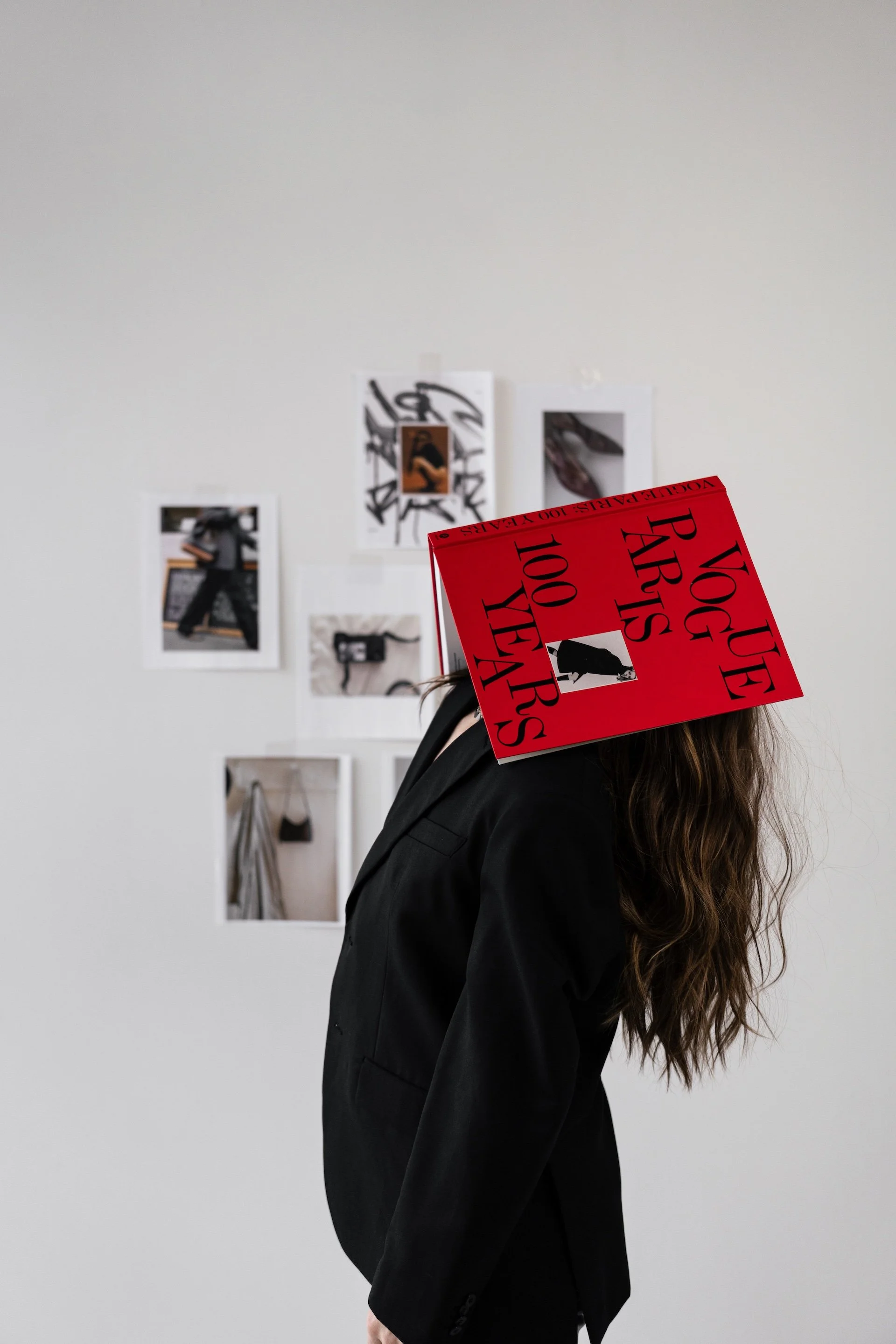 Person with long wavy hair wearing a black suit, her face obscured by a red sign that says 'VOGUE ART'S 100 YEAR'S'. The person is in an art gallery or studio with photos and artwork on a white wall in the background.