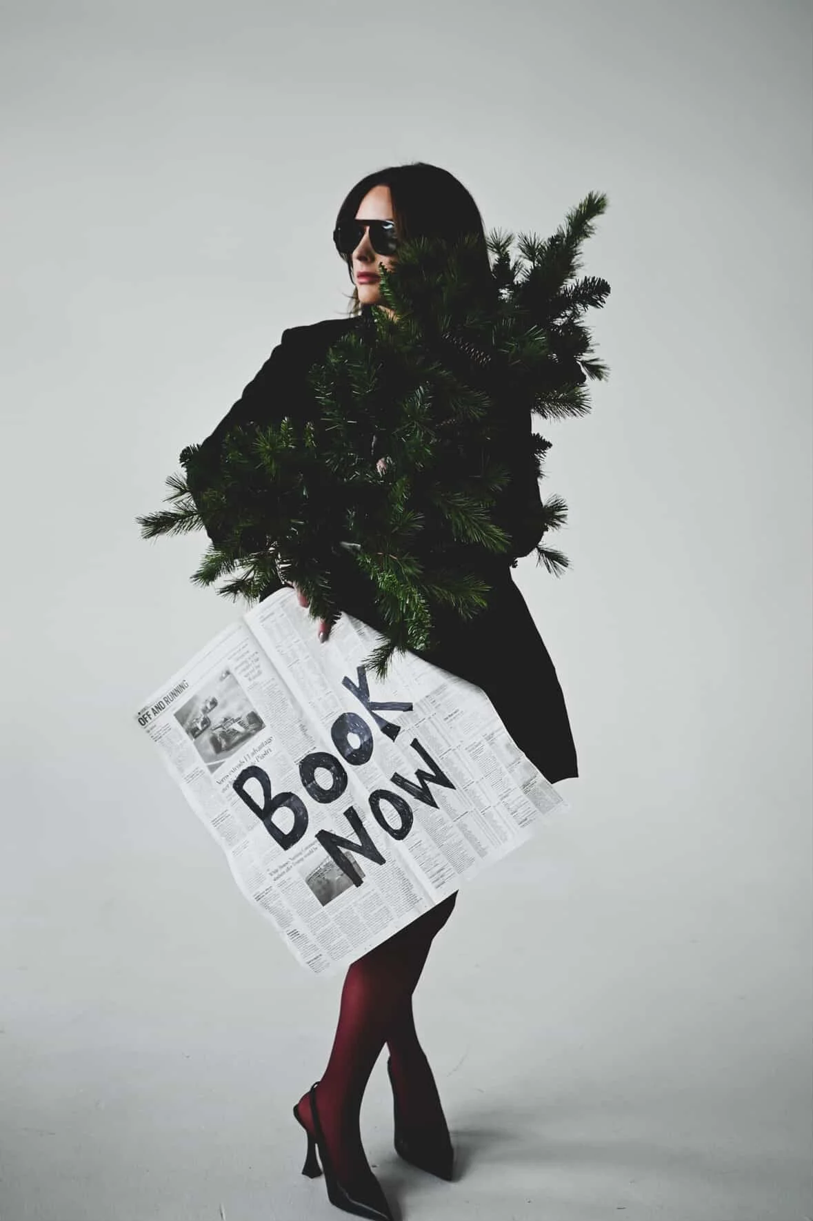A woman dressed in black with black high heels and red stockings, holding a newspaper with the words "BOOK NOW" written on it, and holding a small Christmas tree. She is wearing sunglasses and has shoulder-length dark hair, standing against a plain g