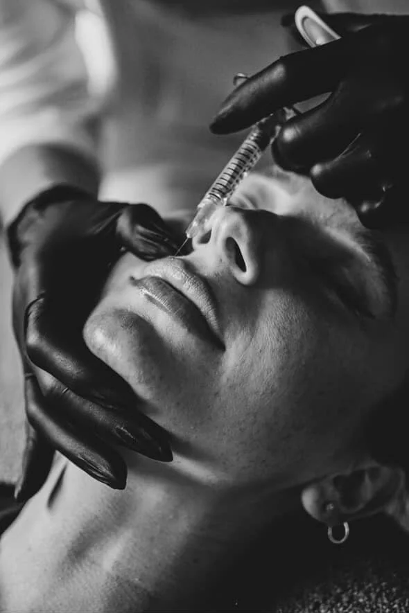 A woman receiving a cosmetic injection in her nose, with a person wearing black gloves administering the injection.