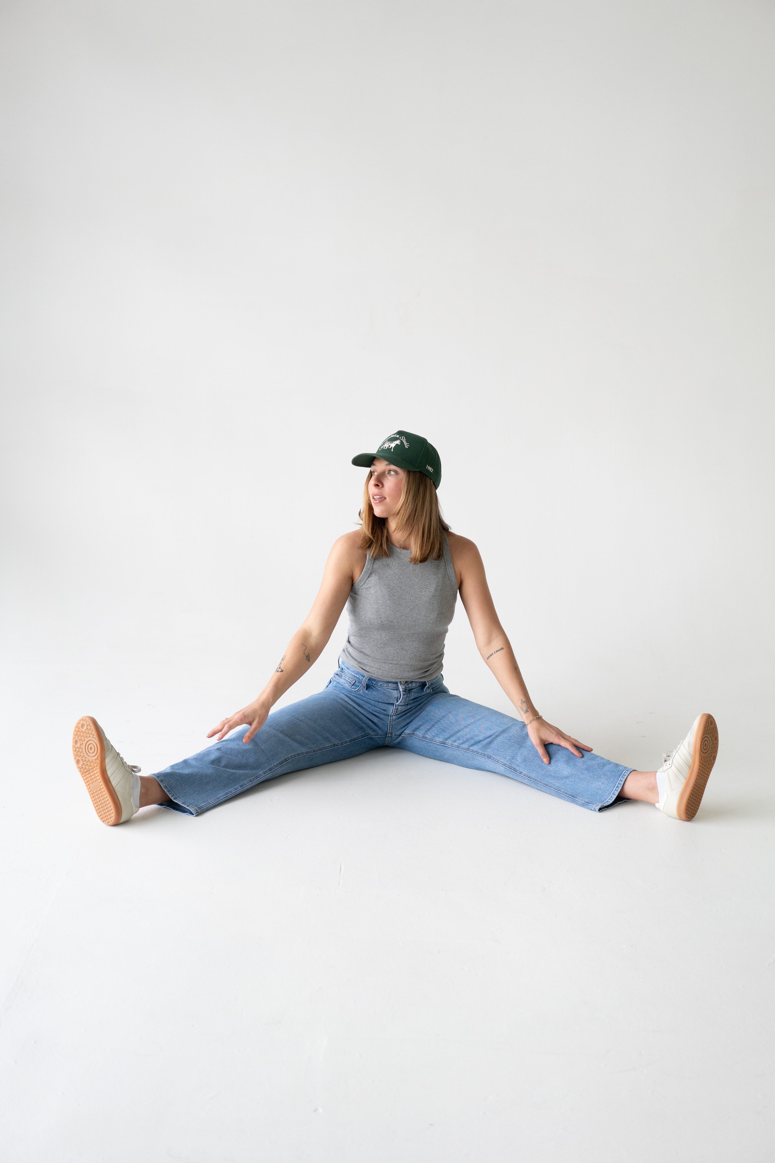 A woman sitting on the floor with legs spread apart, wearing a grey tank top, blue jeans, white sneakers, and a green baseball cap, looking to her left against a plain white background.