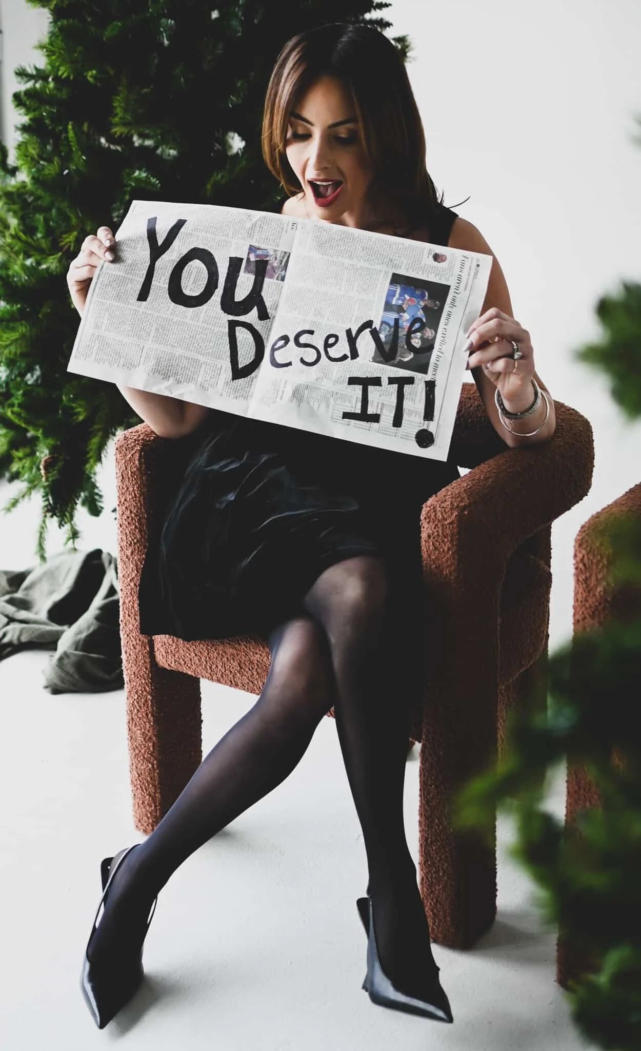 A woman sitting in a chair, holding a newspaper with the words 'You Deserve It!' written on it, with a Christmas tree in the background.