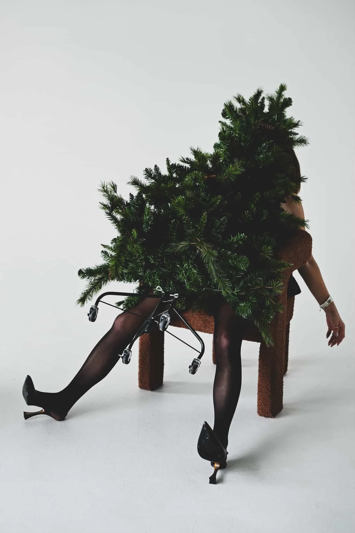 A person sitting on a brown chair with their legs extended forward, wearing black stockings and high heels. They are draped with a Christmas tree covering their upper body and head, with the tree's branches spreading out.