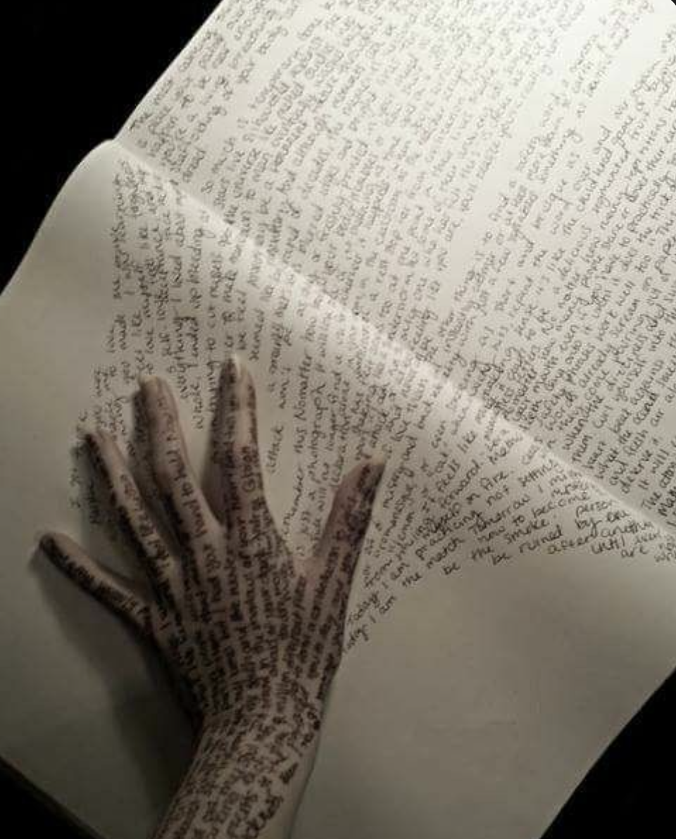 A hand covered with handwritten text, resting on an open notebook or journal filled with handwritten pages.