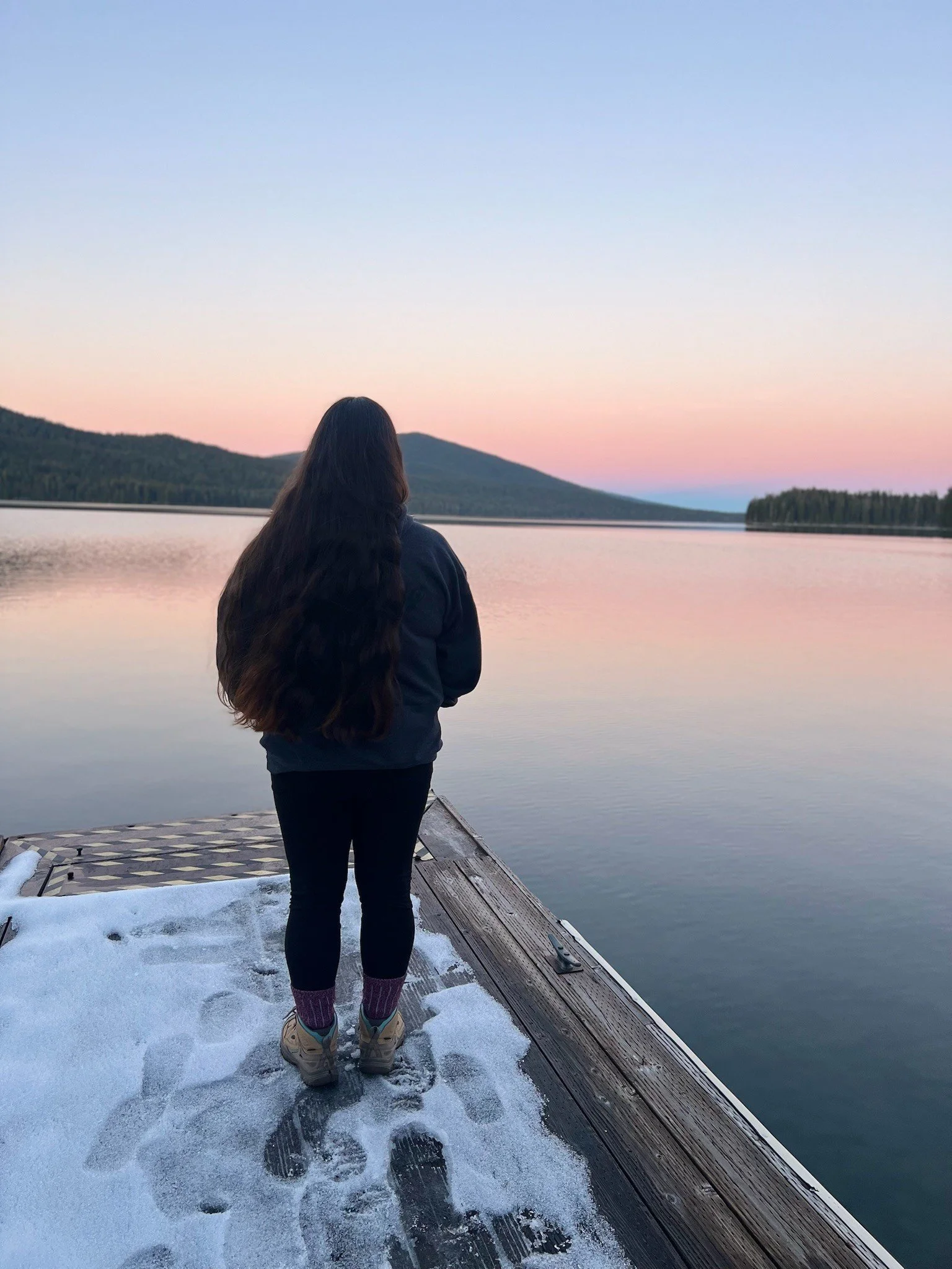 A person with long dark hair, wearing a navy jacket, black pants, and walking boots, stands on a snowy dock facing a calm lake with mountains and a pastel sunset sky in the background.