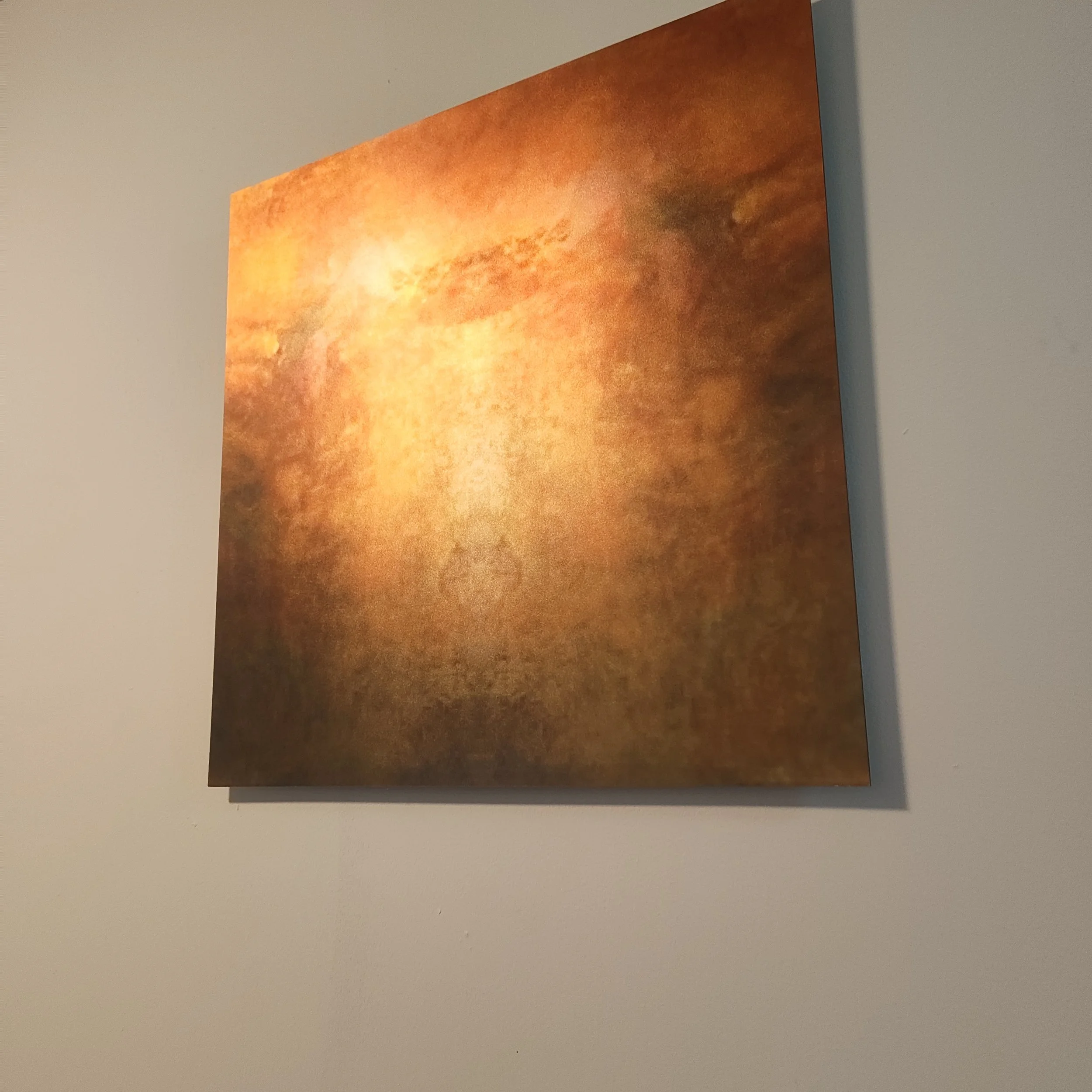 Close-up of a textured painting with warm orange and yellow hues hanging on a white wall.