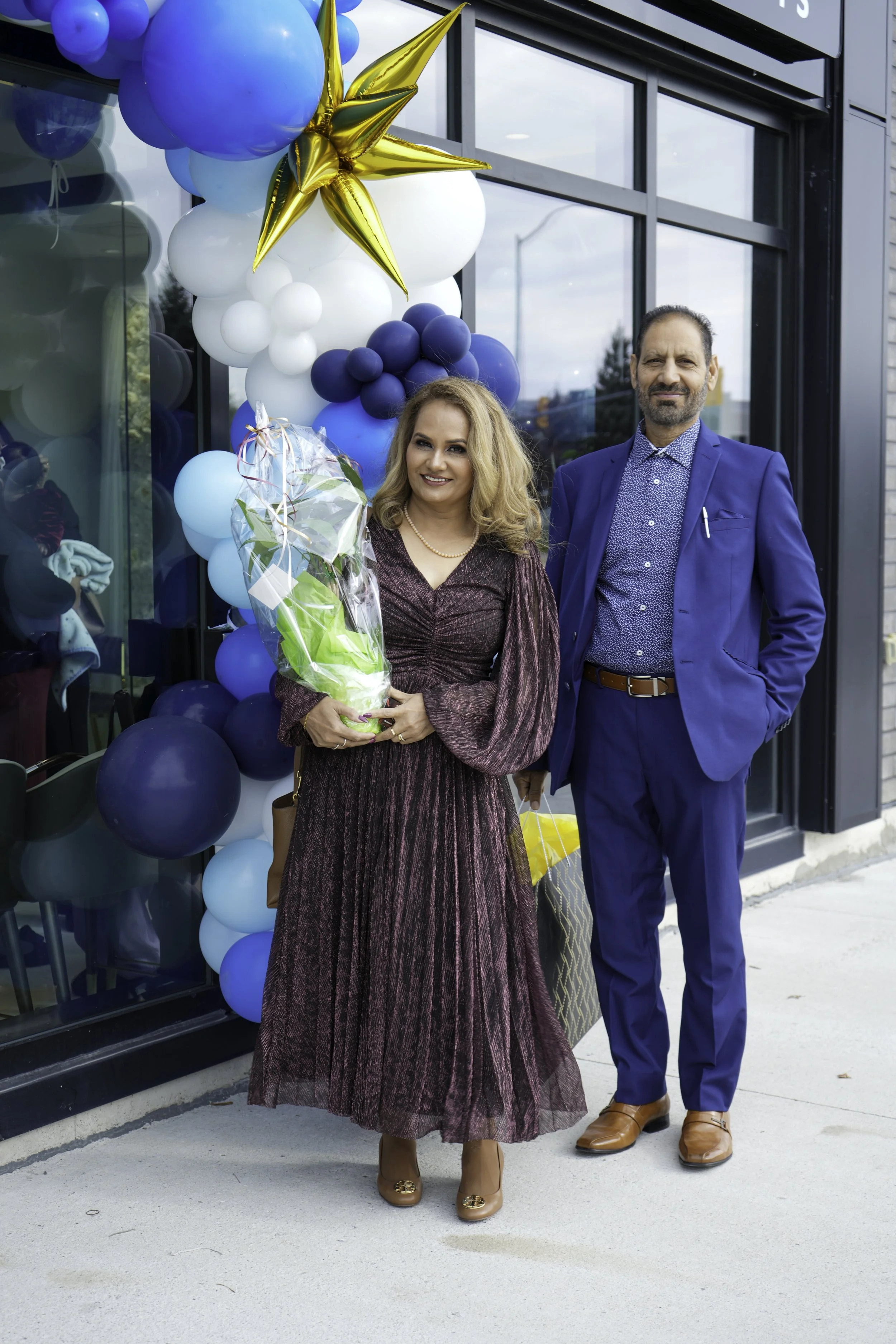Woman and man standing outside decorated with blue, white, and gold balloons, she holding a bouquet of flowers, both dressed in formal attire.