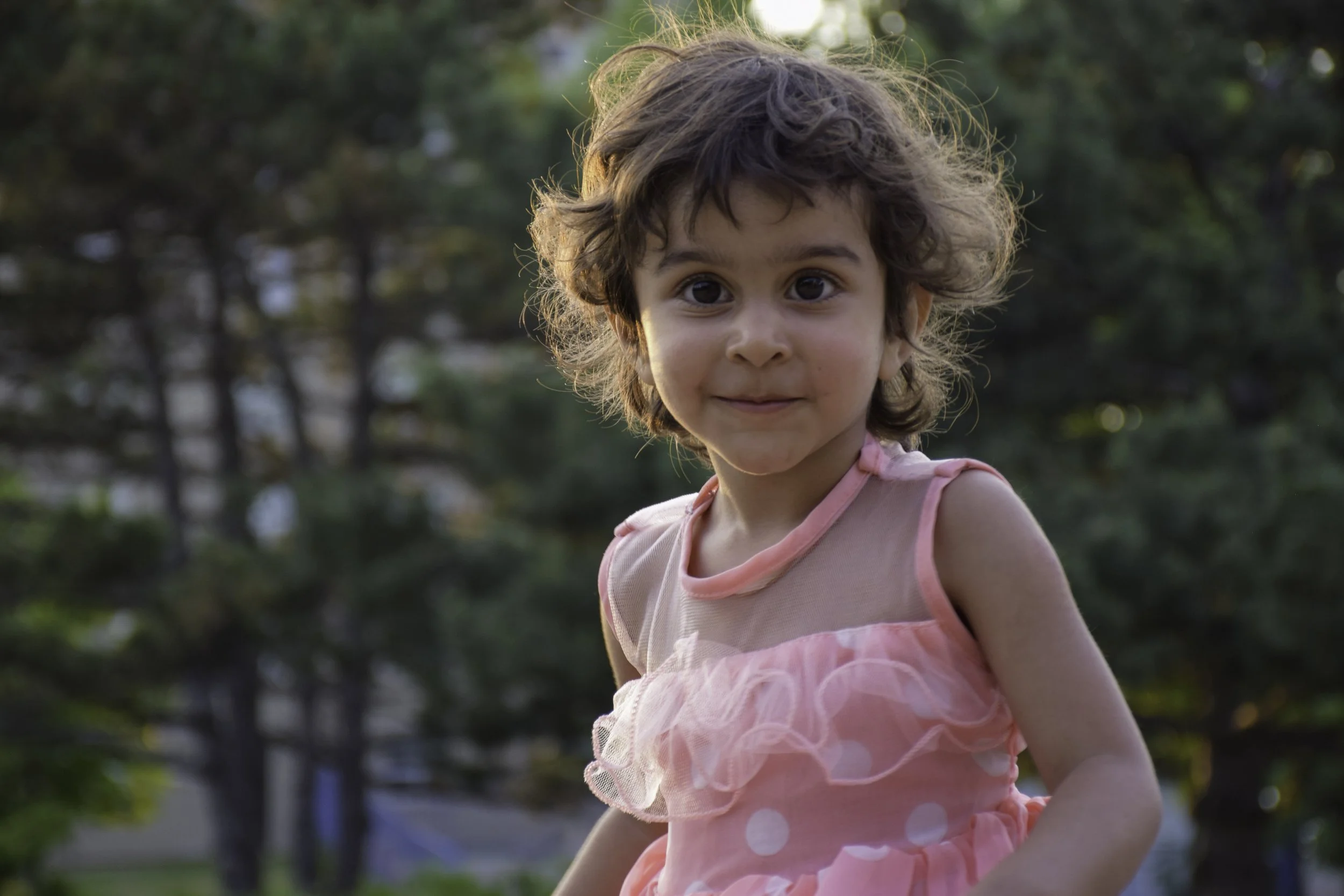A young girl with curly brown hair and big brown eyes wearing a pink dress with white polka dots, outdoors in a park or garden with trees in the background.