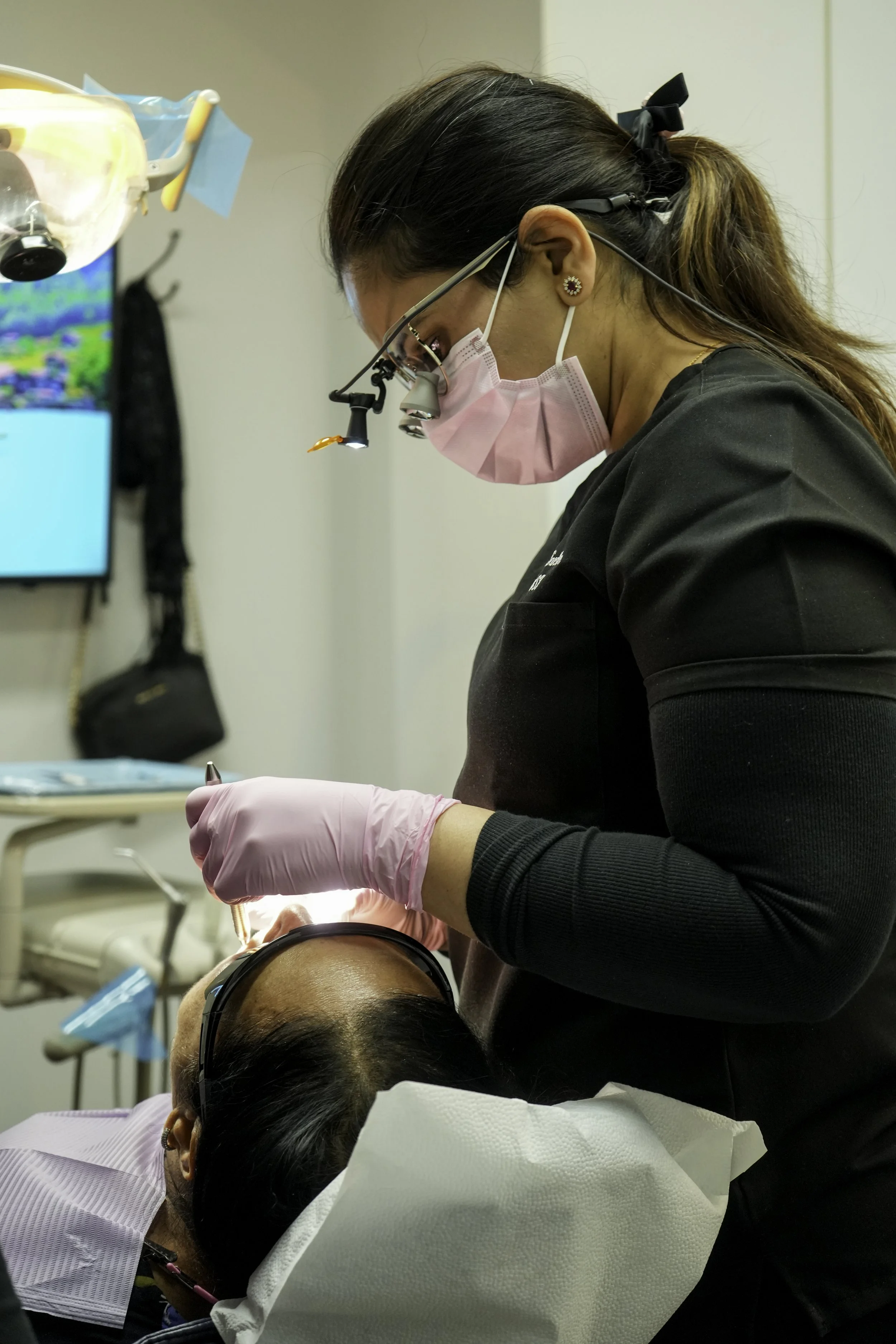 A healthcare professional in scrubs, wearing magnifying glasses with a light, pink mask, and earrings, performing dental or surgical procedure on a patient lying down with protective eyewear, in a clinical setting.