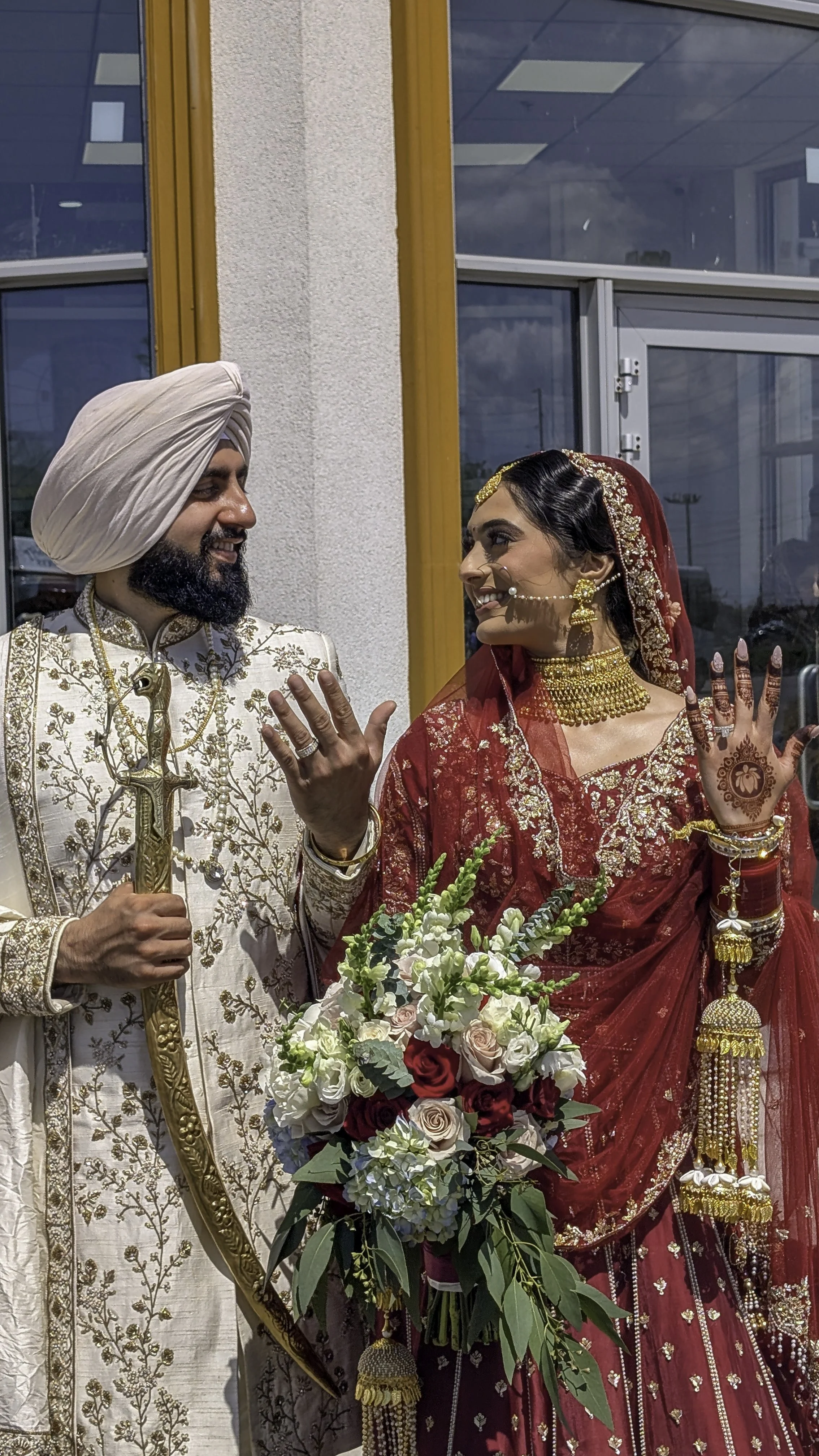 A South Asian bride and groom in traditional wedding attire, smiling and looking at each other, with the bride holding a bouquet of flowers.