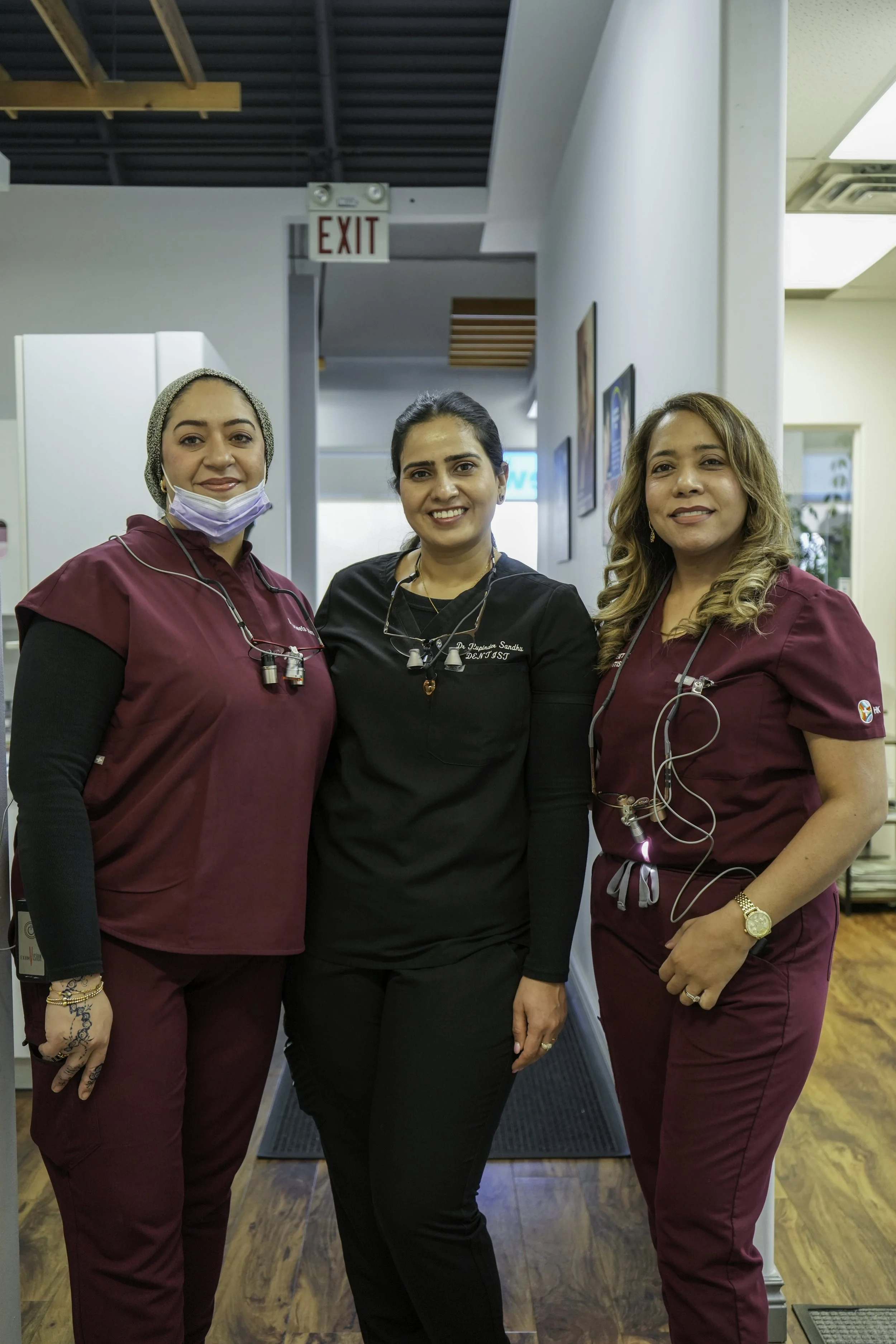 Three women standing together in a professional medical setting. Two women are wearing burgundy scrubs, and one is in black scrubs with the name tag 'Dr. Lipthay Sandhu'. They are smiling, with medical tools around their necks and one wearing a face mask pulled down. The background features white walls, framed artwork, and a hallway leading to an exit.