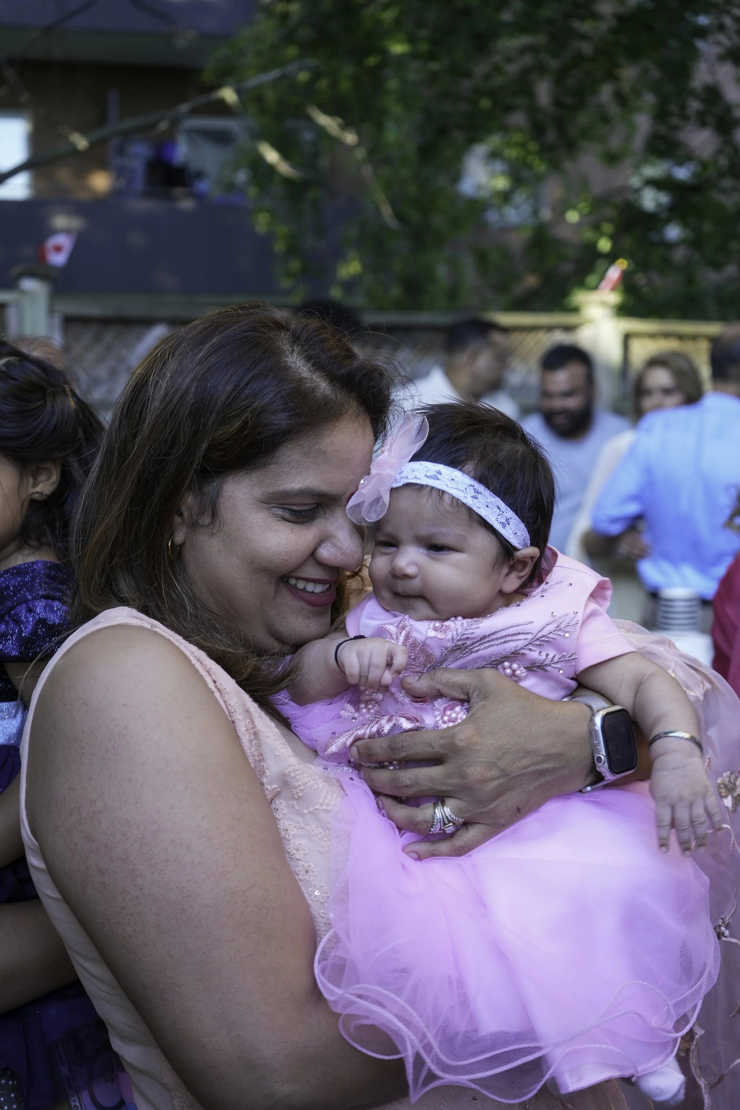 A woman holding a baby girl dressed in a pink dress and headband, smiling at each other at an outdoor gathering with other people in the background.
