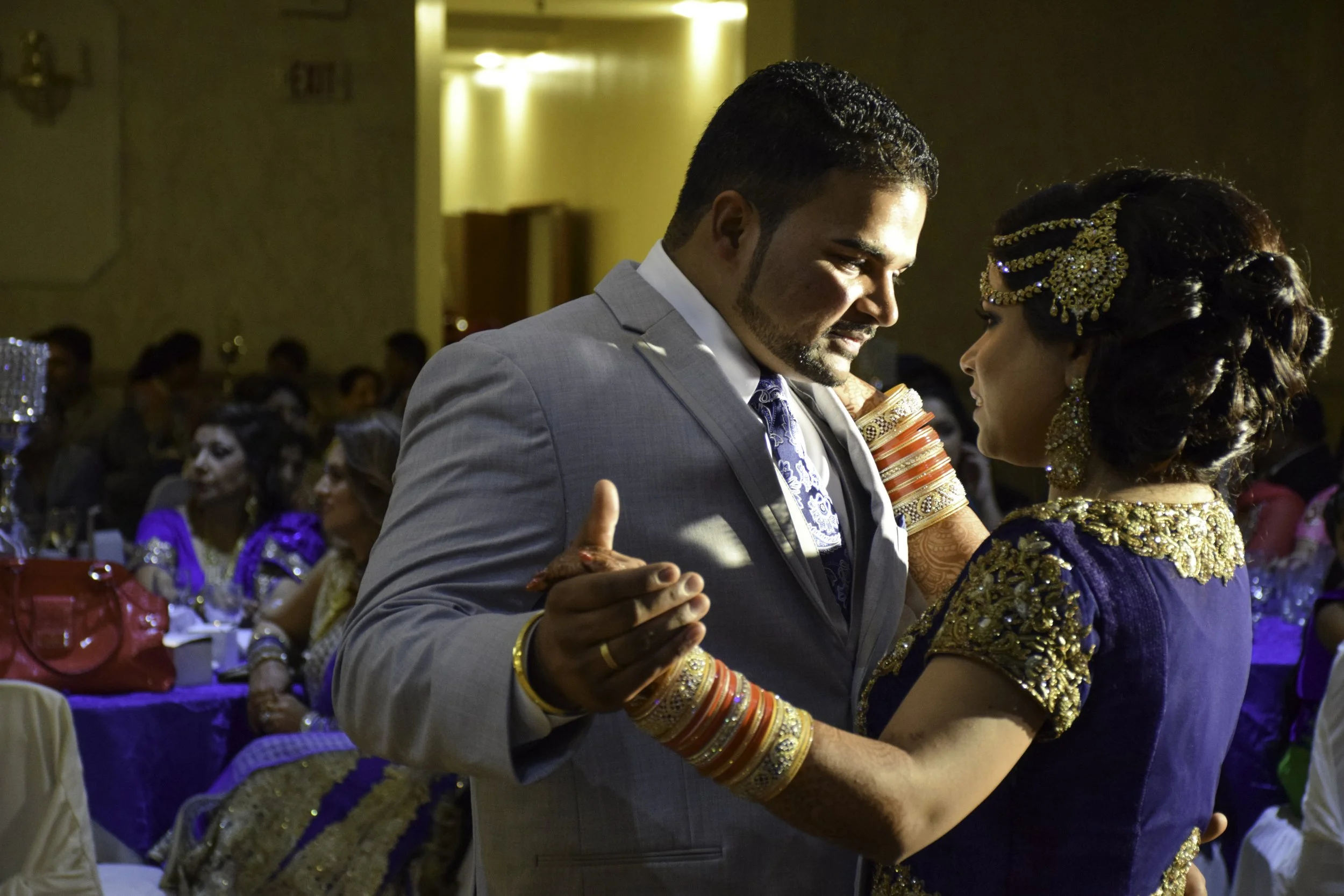 A couple dancing at a wedding reception, dressed in formal attire with the woman wearing traditional Indian jewelry and the man in a light grey suit with a blue patterned tie.