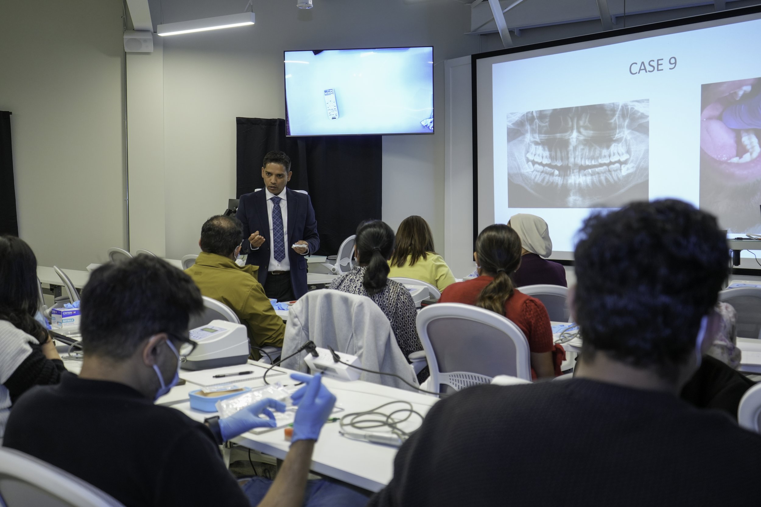 A professional presenter in a suit stands in front of an audience during a conference or seminar. The audience is seated at tables with writing materials and medical gloves, and some participants are wearing masks. There is a large screen displaying a presentation slide with the text "CASE 9" and an X-ray of teeth. Another smaller screen shows a close-up of a mobile phone.