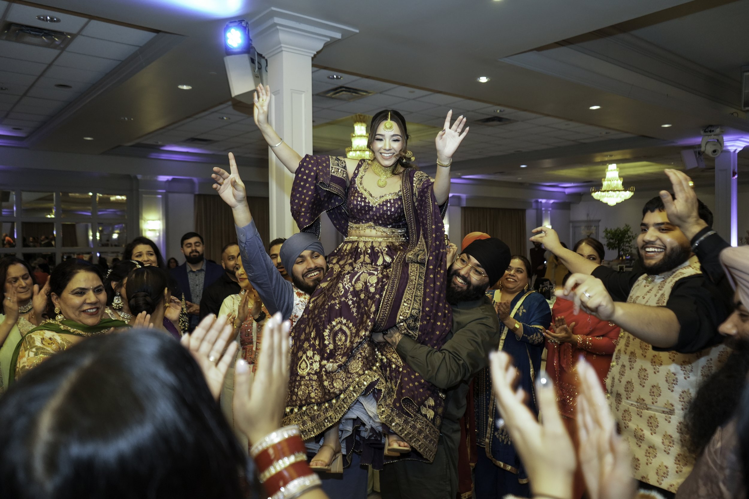 A woman dressed in traditional Indian attire smiling joyfully while being lifted by a man during a celebration, surrounded by other cheerful guests.