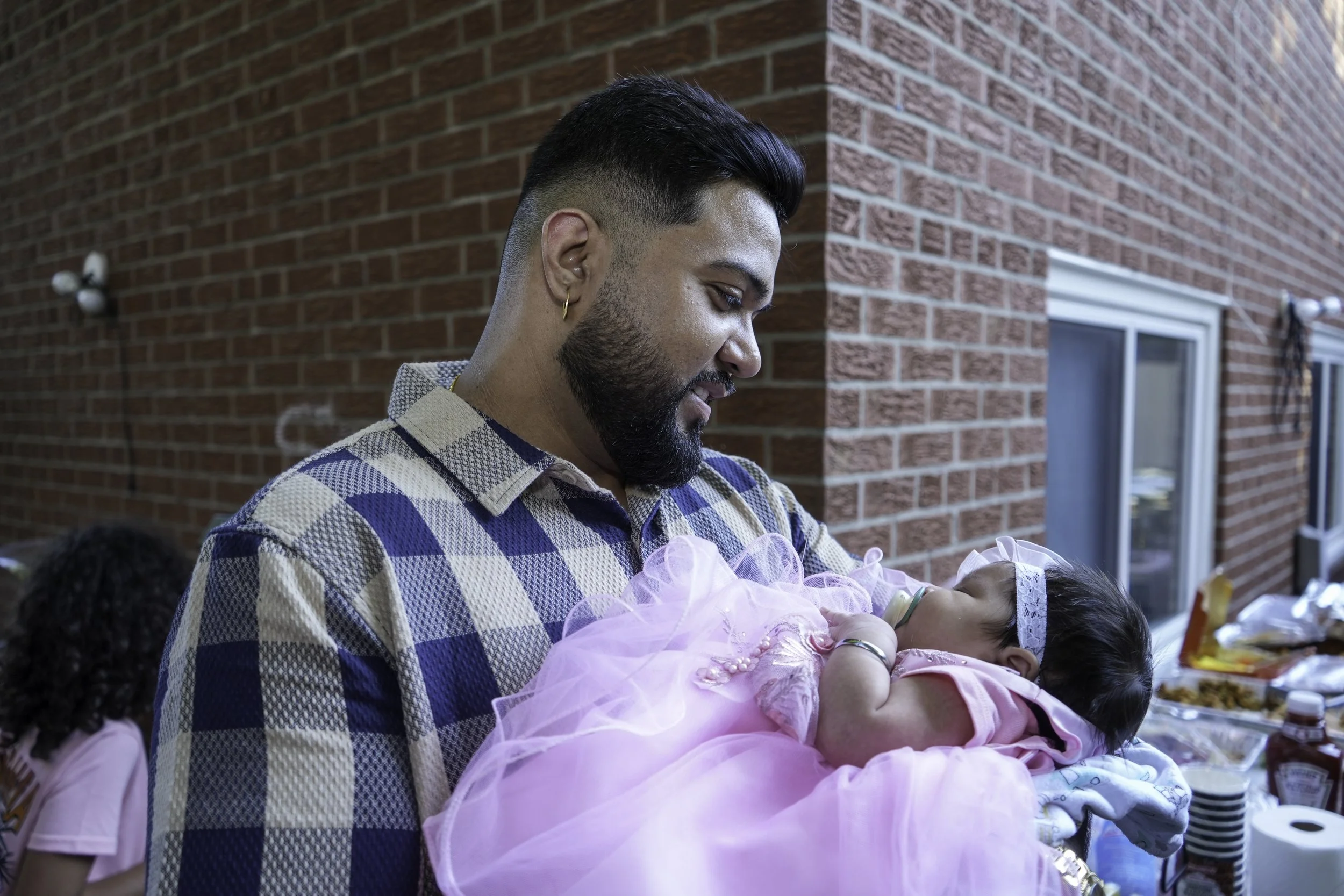 A man with dark hair, a beard, and gold hoop earrings is smiling while holding a young girl dressed in a pink tutu at an outdoor gathering against a brick wall.