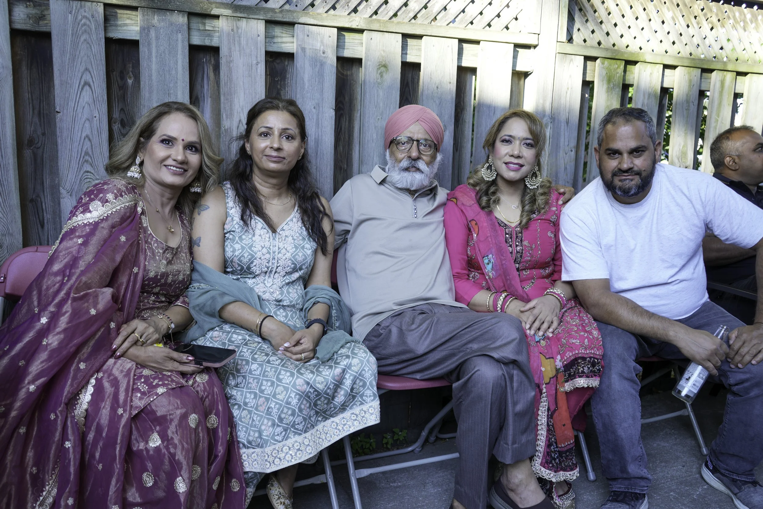 A group of six people sitting outdoors in front of a wooden fence, smiling and posing for the photo. The group includes women dressed in traditional Indian attire and men, one wearing a pink turban and glasses.