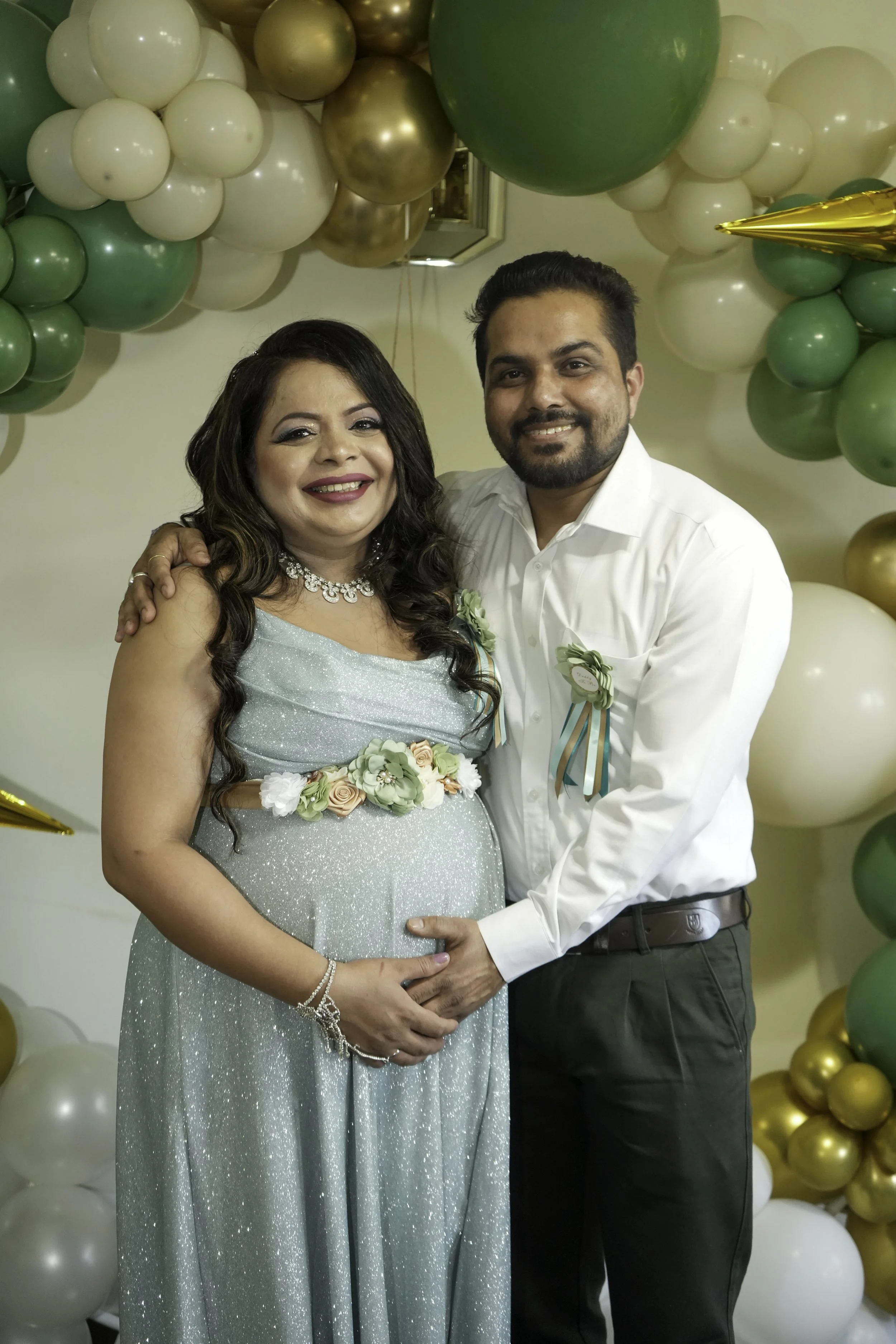 A smiling woman in a light blue, glittery dress holding her pregnant belly stands next to a smiling man in a white shirt. They are in front of a backdrop of green, white, gold, and silver balloons, celebrating a special occasion.