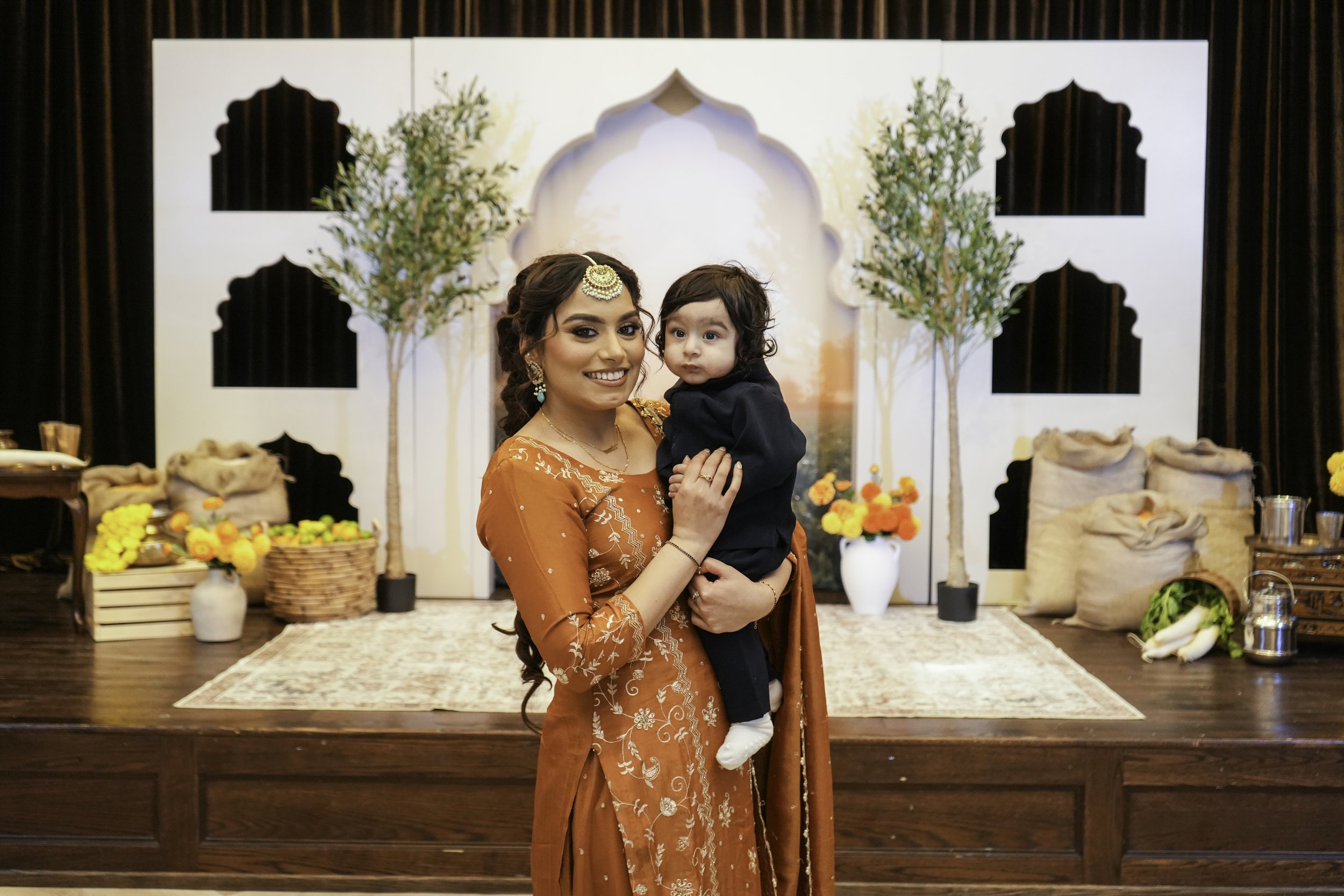 A woman in traditional Indian attire holding a young child at a celebration or event with decorated backdrop, flowers, and items on a table.
