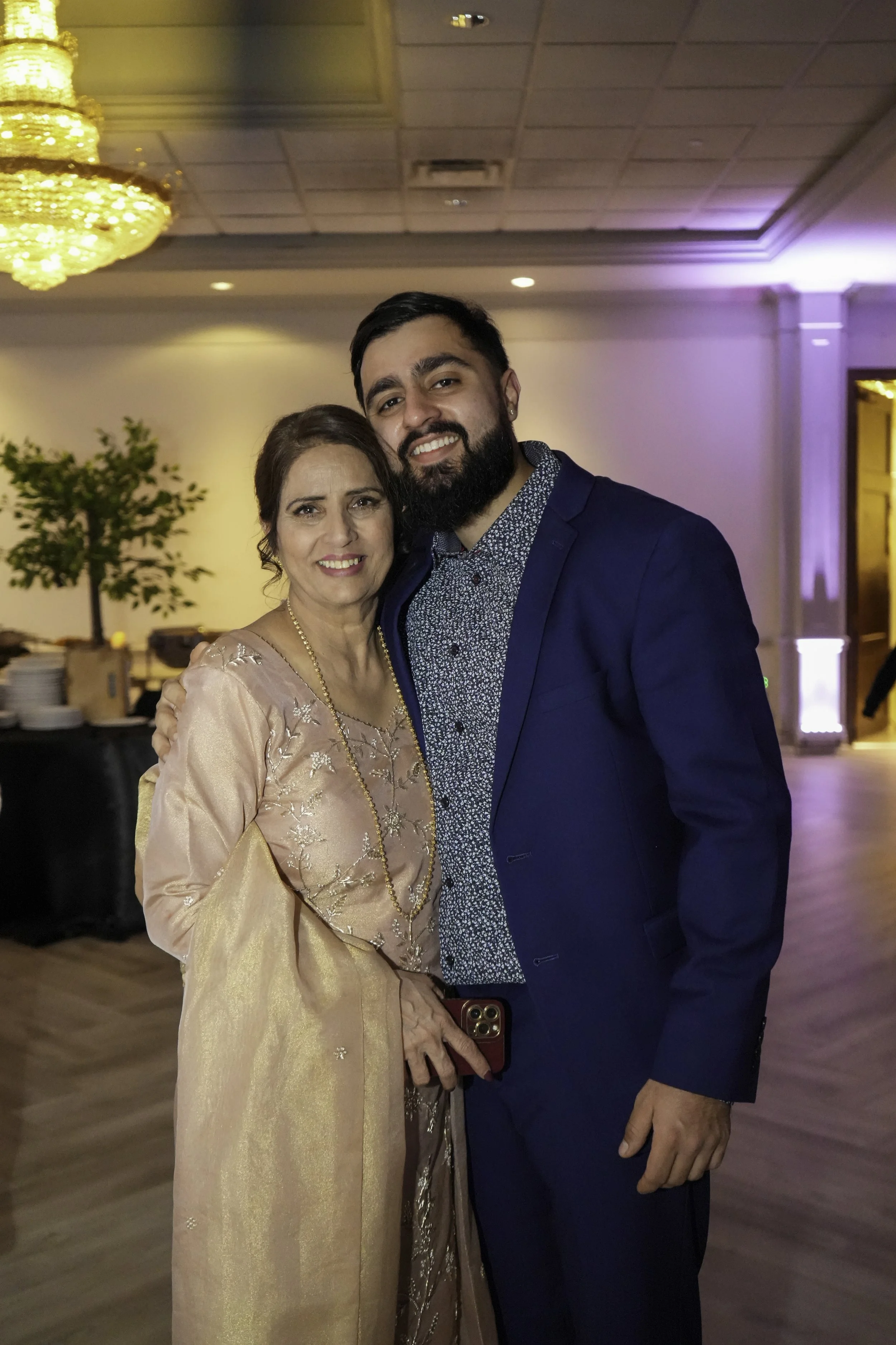 A woman and a man smiling, embracing each other at a social event or celebration in a decorated room with a chandelier, potted plant, and buffet tables in the background.