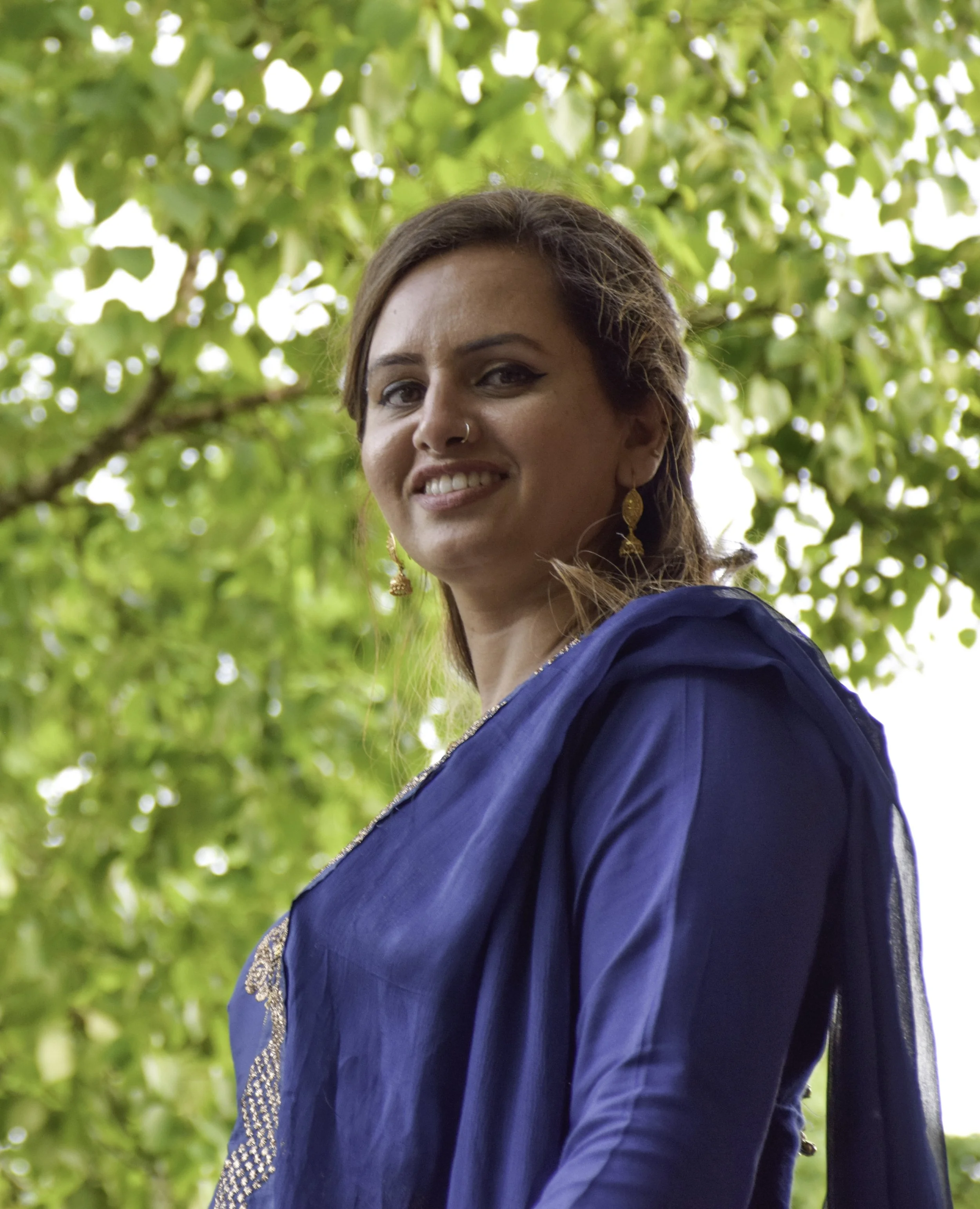 A woman with brown hair, earrings, and a nose ring smiling outdoors among green leaves, wearing a blue dress or blouse.