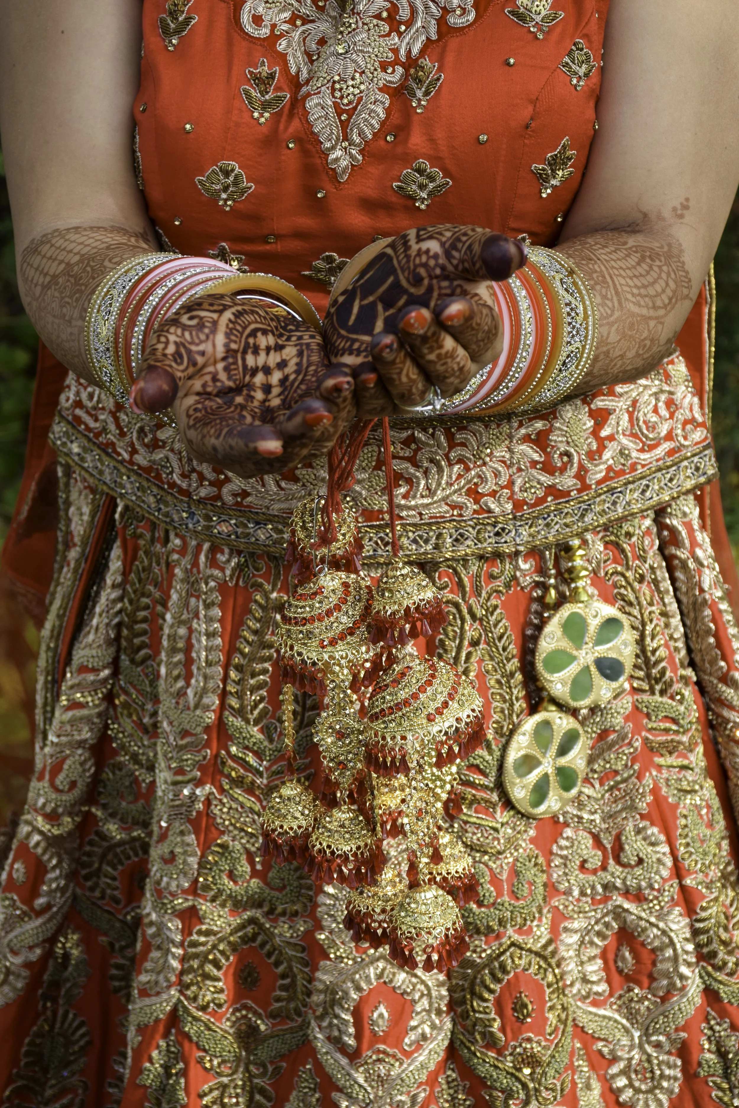 Close-up of a person wearing traditional Indian attire, including a richly embroidered red and gold dress and elaborate bangles, with henna designs on their hands.