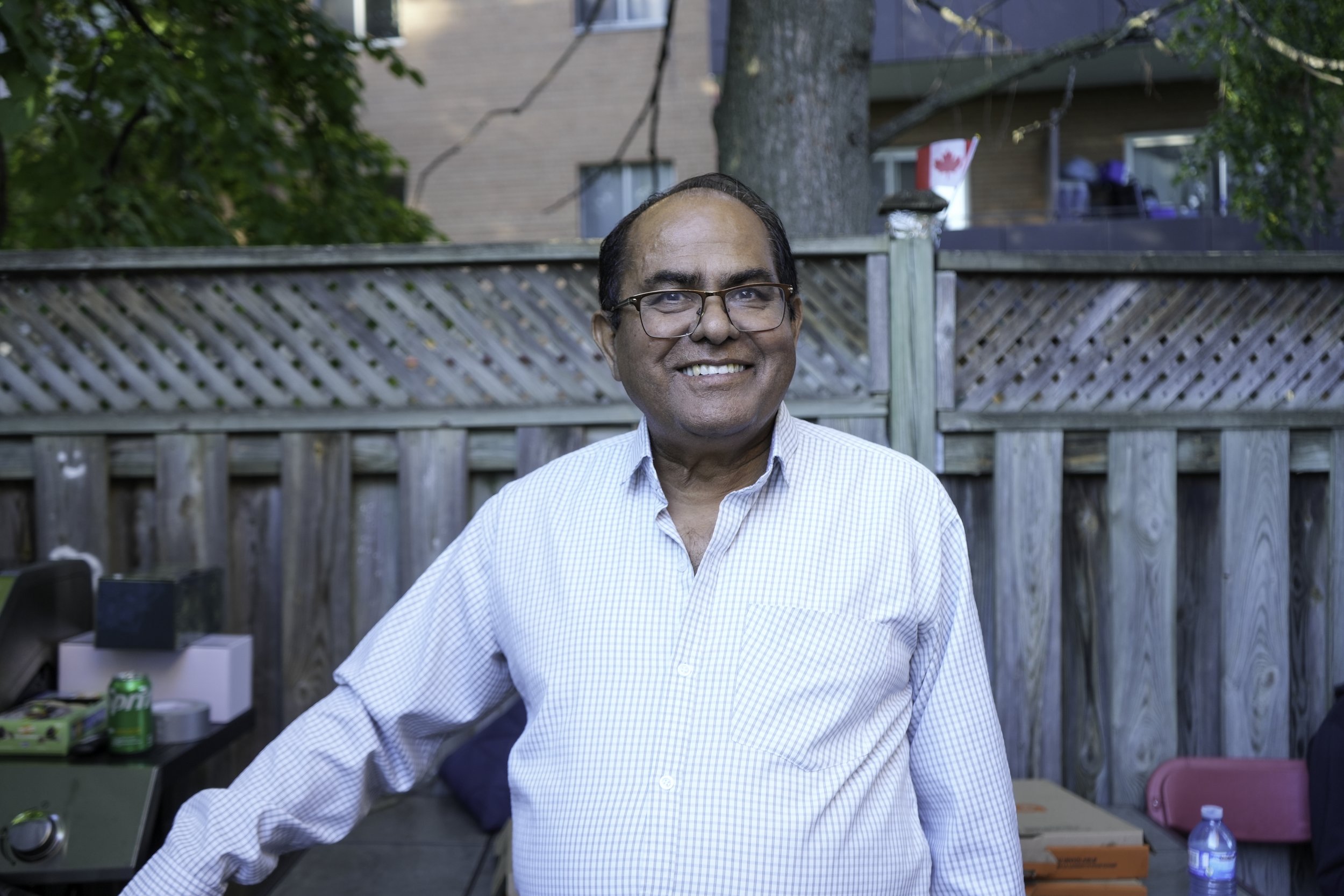 Smiling man with glasses wearing a light blue checkered shirt standing outdoors in a backyard with a wooden fence, trees, and a Canadian flag on a house in the background.