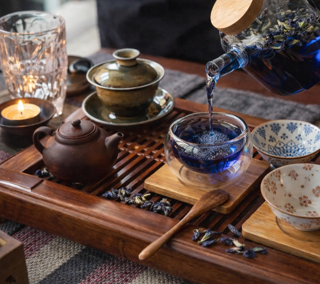 Pouring blue herbal tea into a glass teacup on a wooden tray, with teapots, bowls, a small candle, and a glass of water nearby on a table.