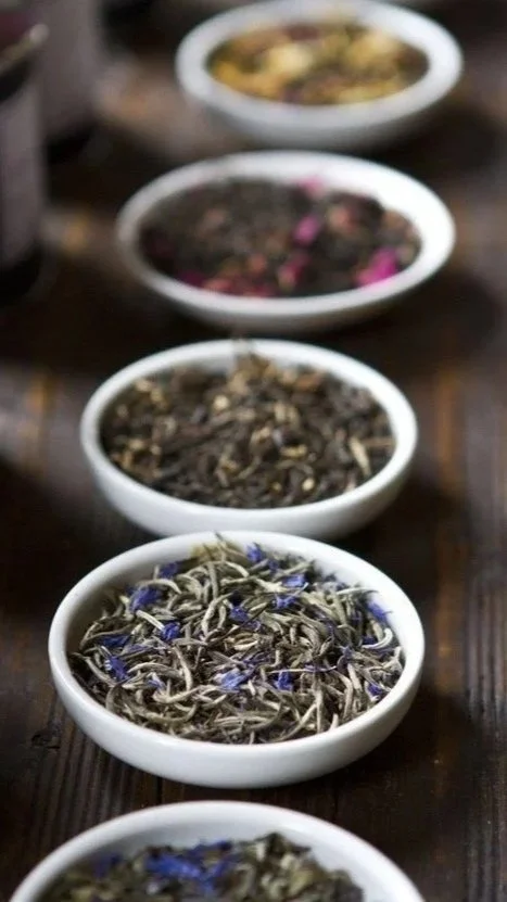 Several small white bowls filled with different dried herbs or flowers, arranged in a line on a wooden table.