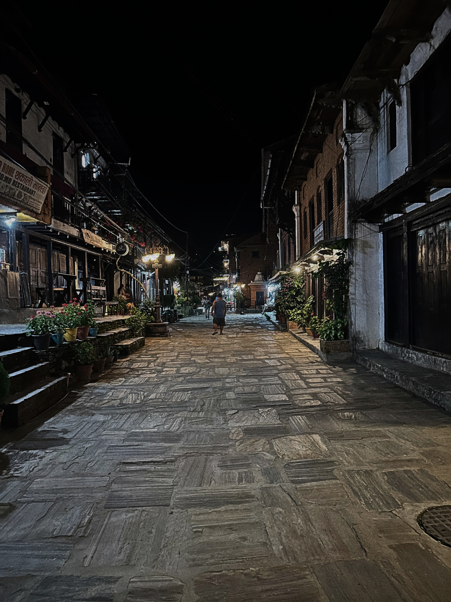 A quiet, paved street at night with buildings on both sides, some with decorative plants and lights. A person is walking down the street, and the sky is dark.