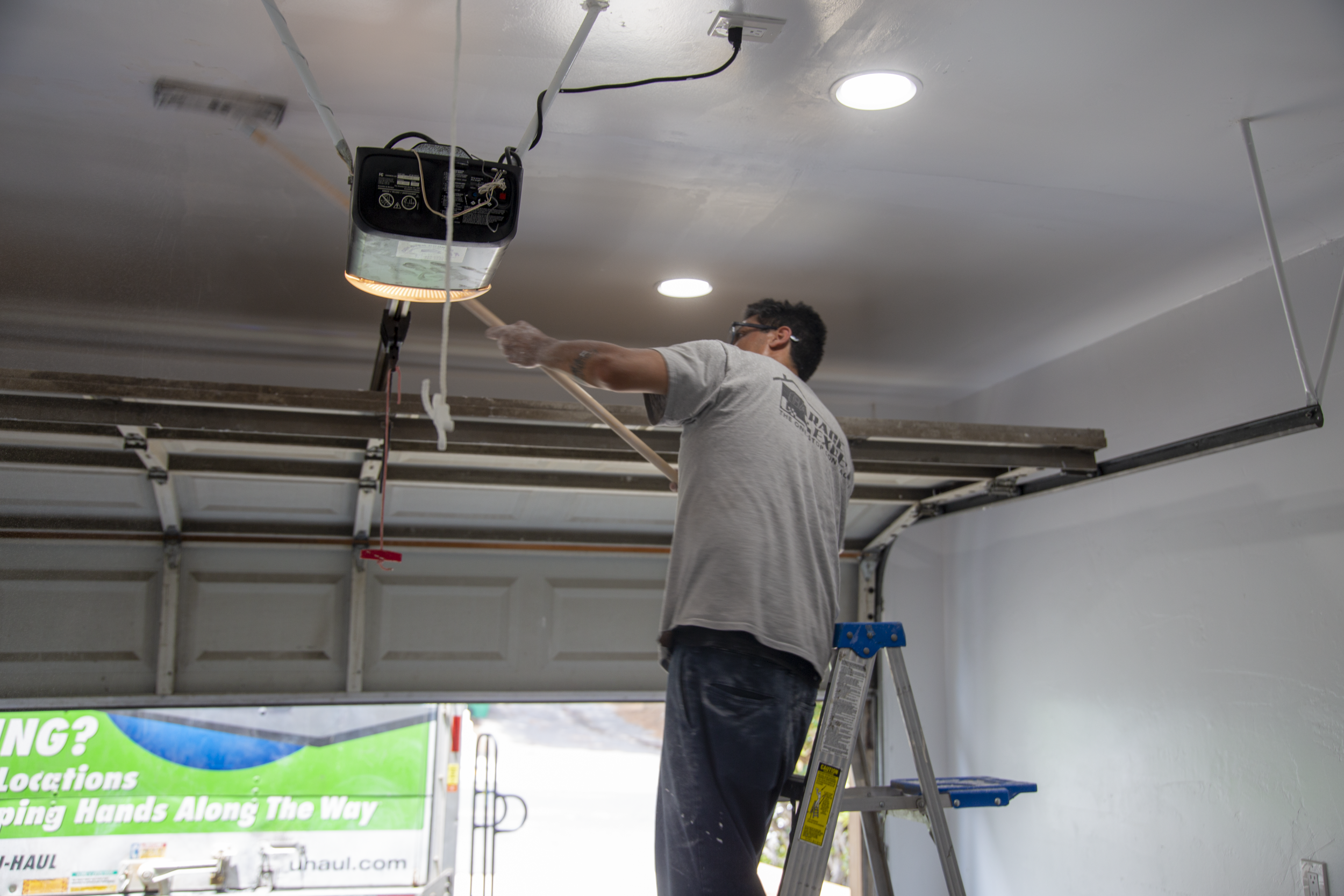 A man standing on a ladder using a long tool to work on the garage ceiling near a mounted garage door opener.