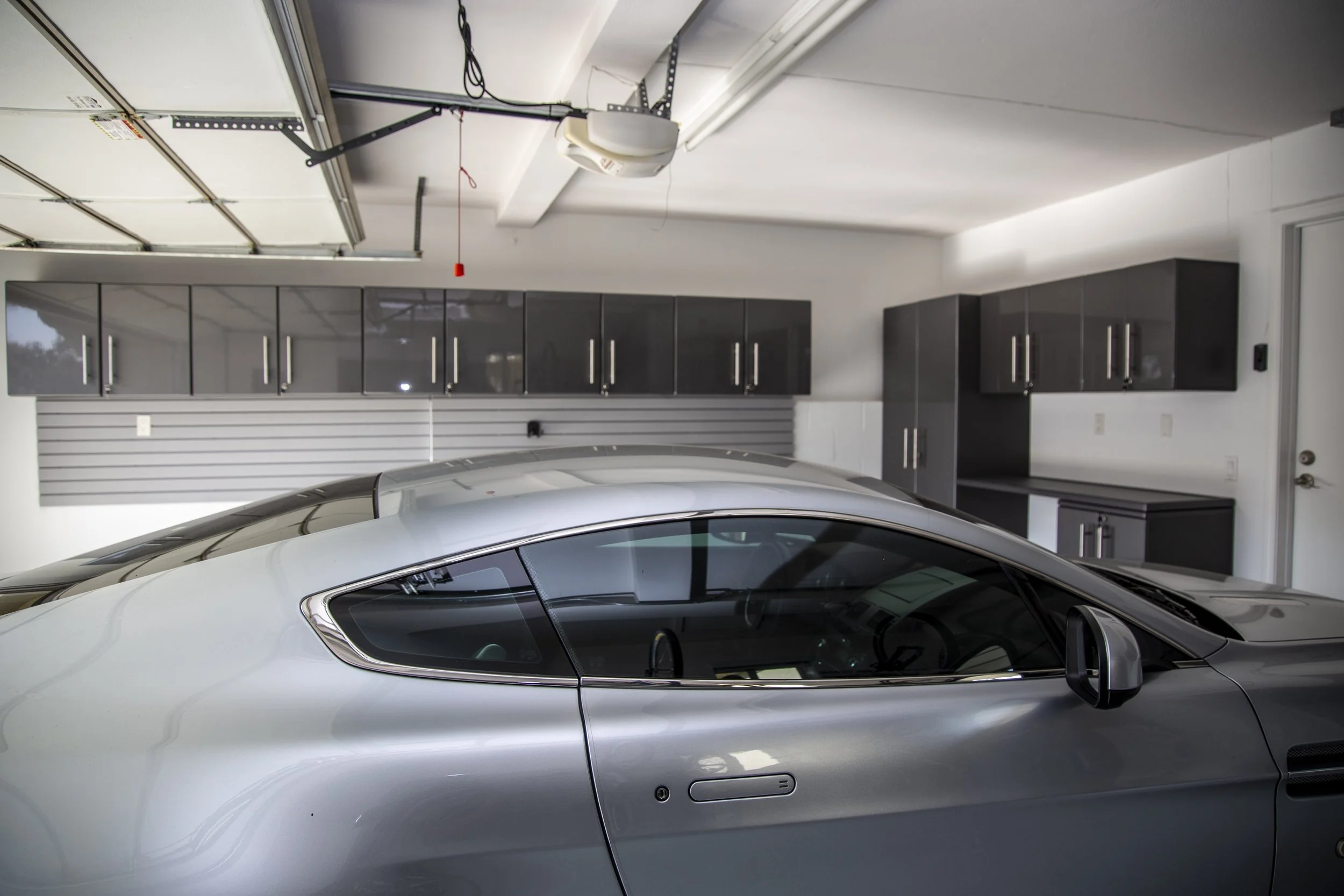 Inside a garage with a silver sports car parked, gray cabinets on the wall, and garage door mechanisms visible on the ceiling.