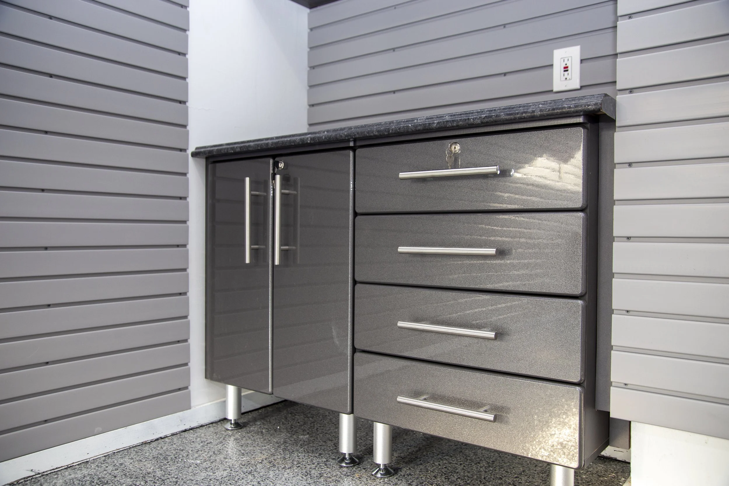 A modern kitchen cabinet with three drawers and a double-door compartment, stainless steel handles, set under a black granite countertop, against gray paneled walls, with an electrical outlet above.