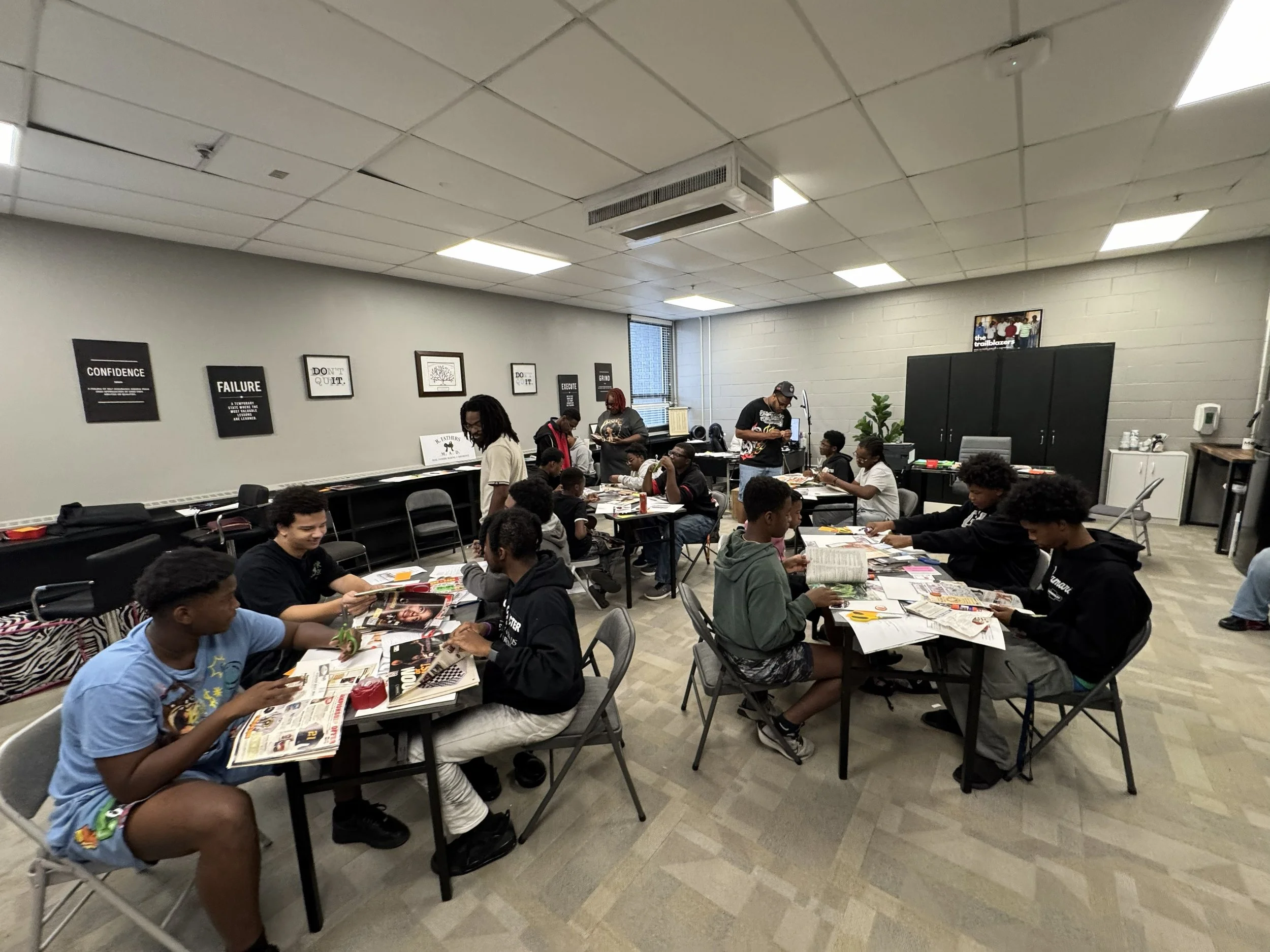 A group of children and teenagers sitting at tables in a classroom or community room, engaging with magazines and papers. There are a few adults standing, interacting with the group. The room has white walls, framed quotes, and a large window in the 