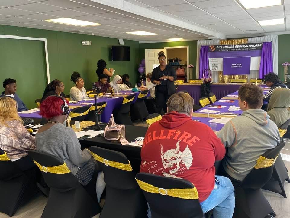A group of people attending a seminar or workshop in a decorated conference room with purple and gold accents. A woman stands at the front speaking to the audience.