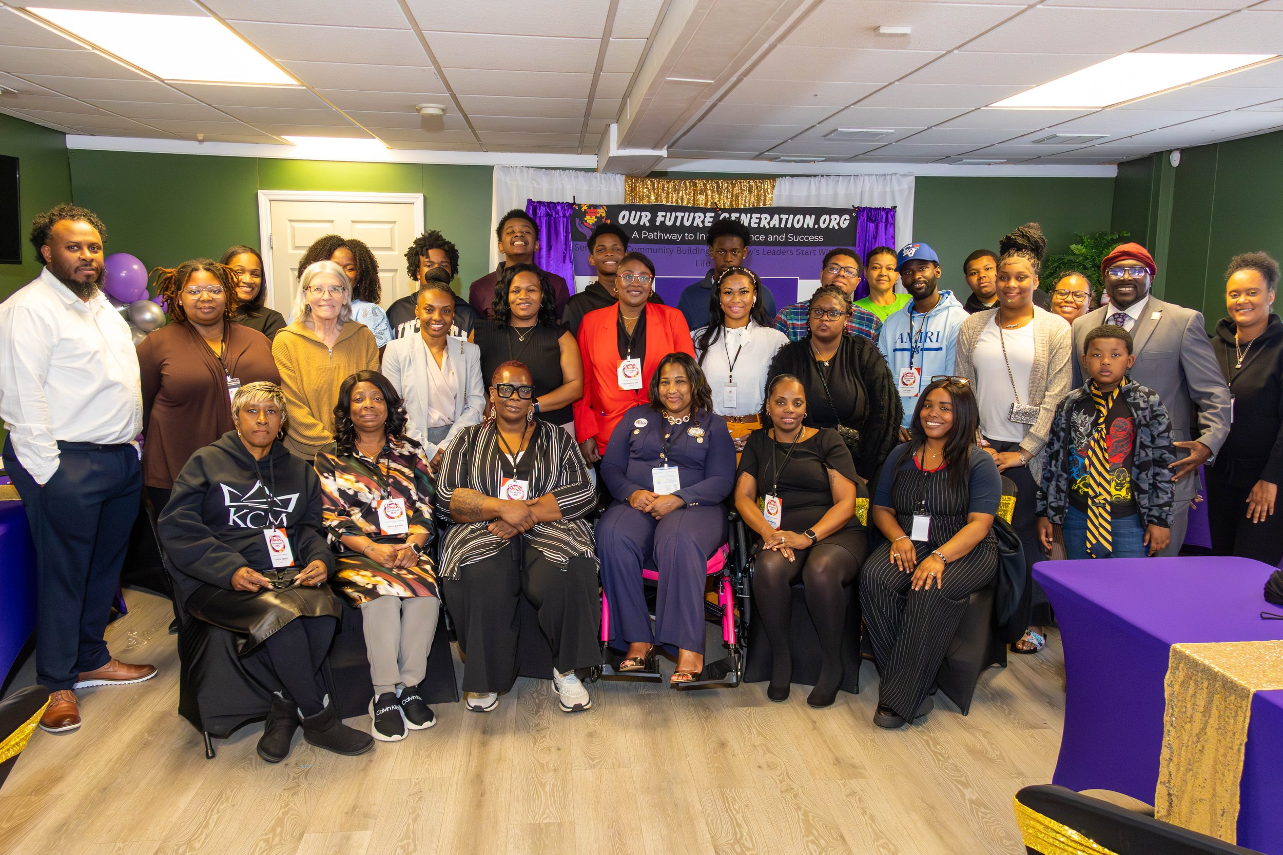Group photo of diverse individuals, including women, men, and children, at a community event in a room decorated with purple and gold with balloons and a backdrop that says "Our Future Generation.org".