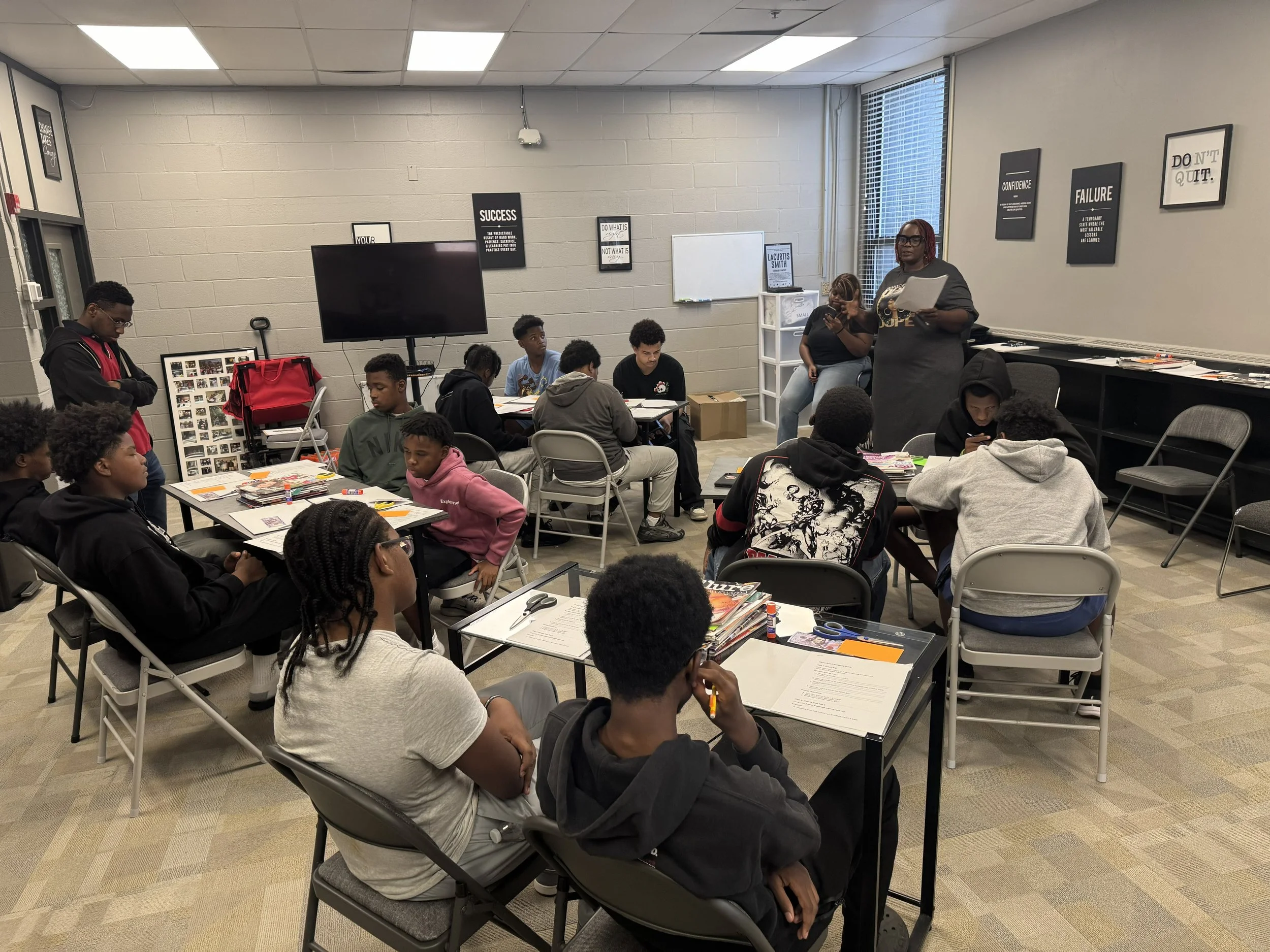 A classroom with students sitting at tables listening to a woman standing at the front holding papers. The classroom walls have motivational posters and a whiteboard. A large monitor is mounted on the wall.