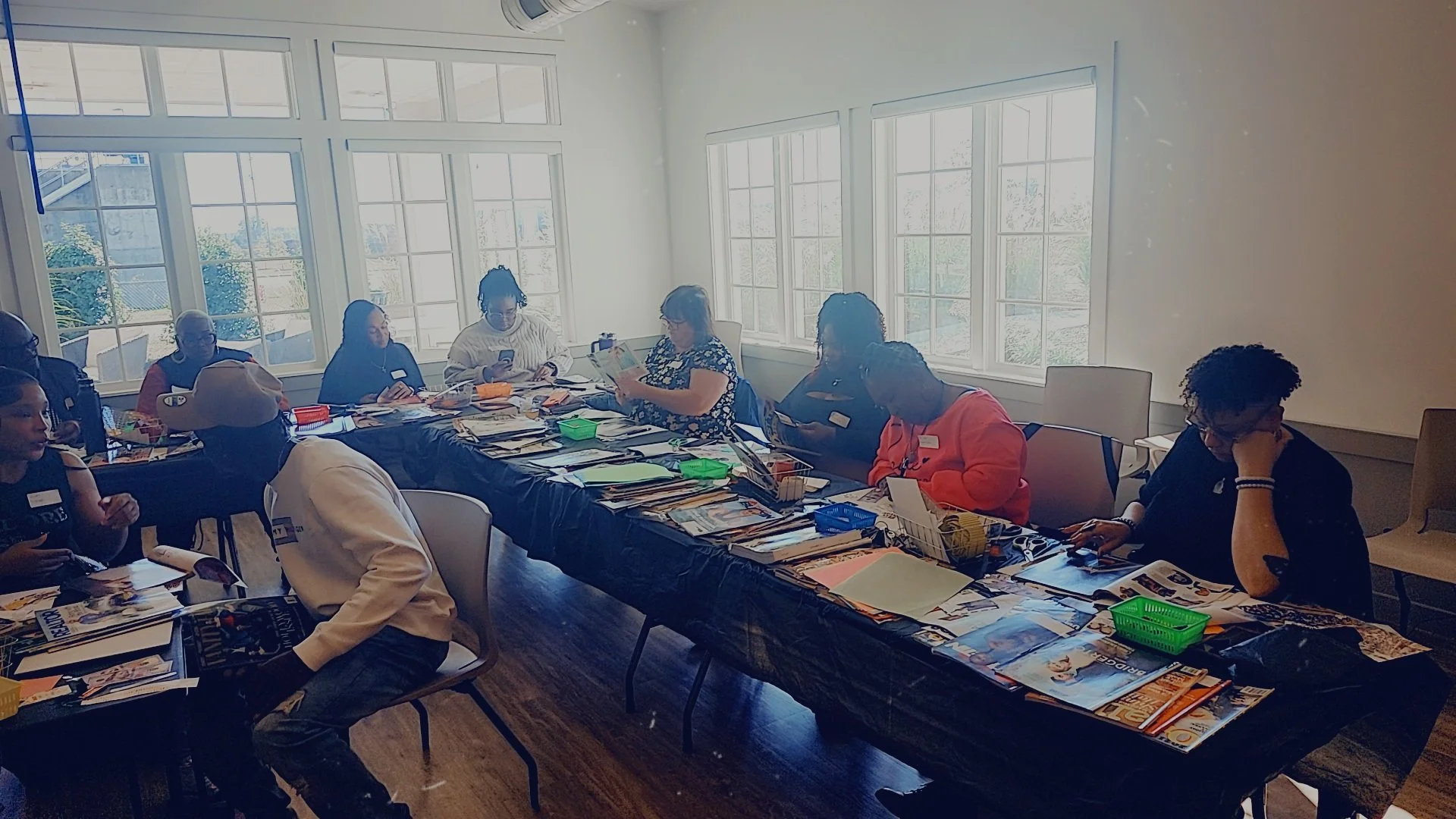 A group of people sitting around a long table in a bright room with large windows, engaged in reading or working with various materials and magazines.