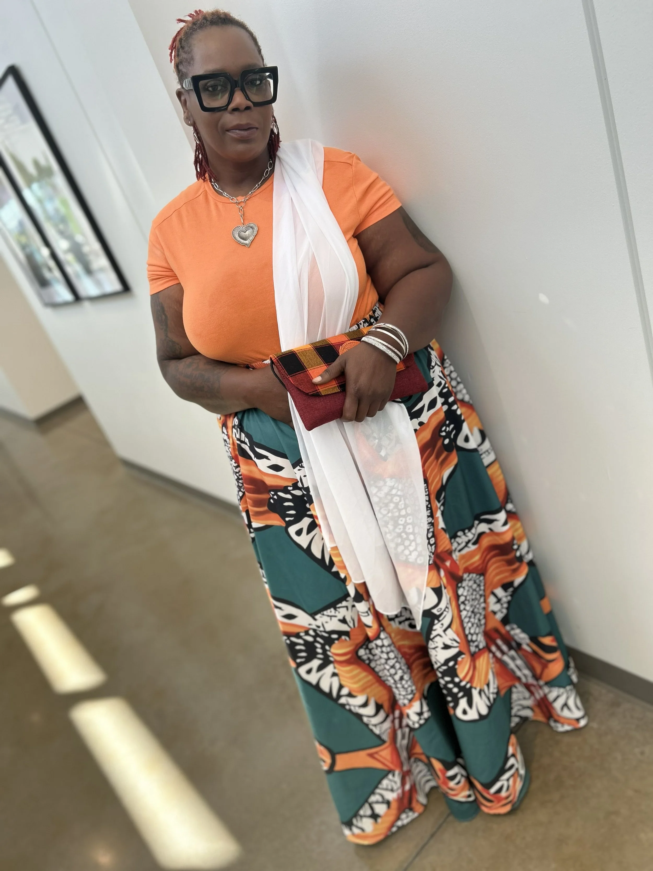 A woman with red hair styled in dreadlocks, wearing large black glasses, a silver chain necklace with a heart pendant, an orange shirt, and a patterned skirt, stands against a white wall holding a folded cloth with red, black, yellow, and green colors.