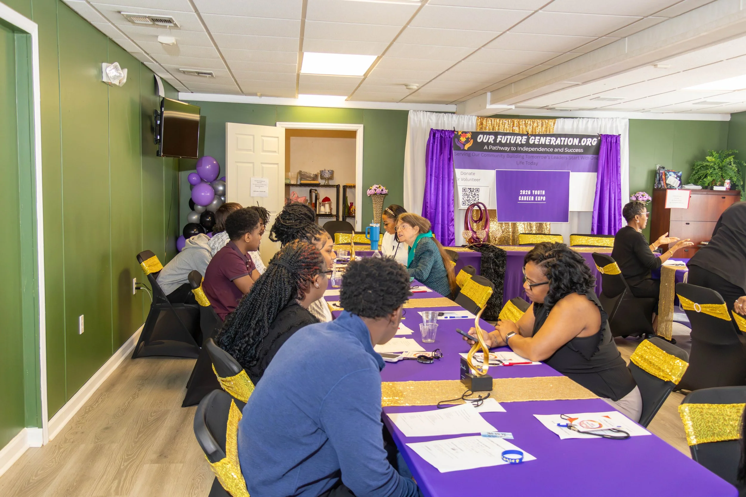 A group of people, mostly young adults, sitting at a long table with purple tablecloths, attending a community event. There are balloons, a banner reading 'OUR FUTURE GENERATION.ORG', and various decorations in the background.