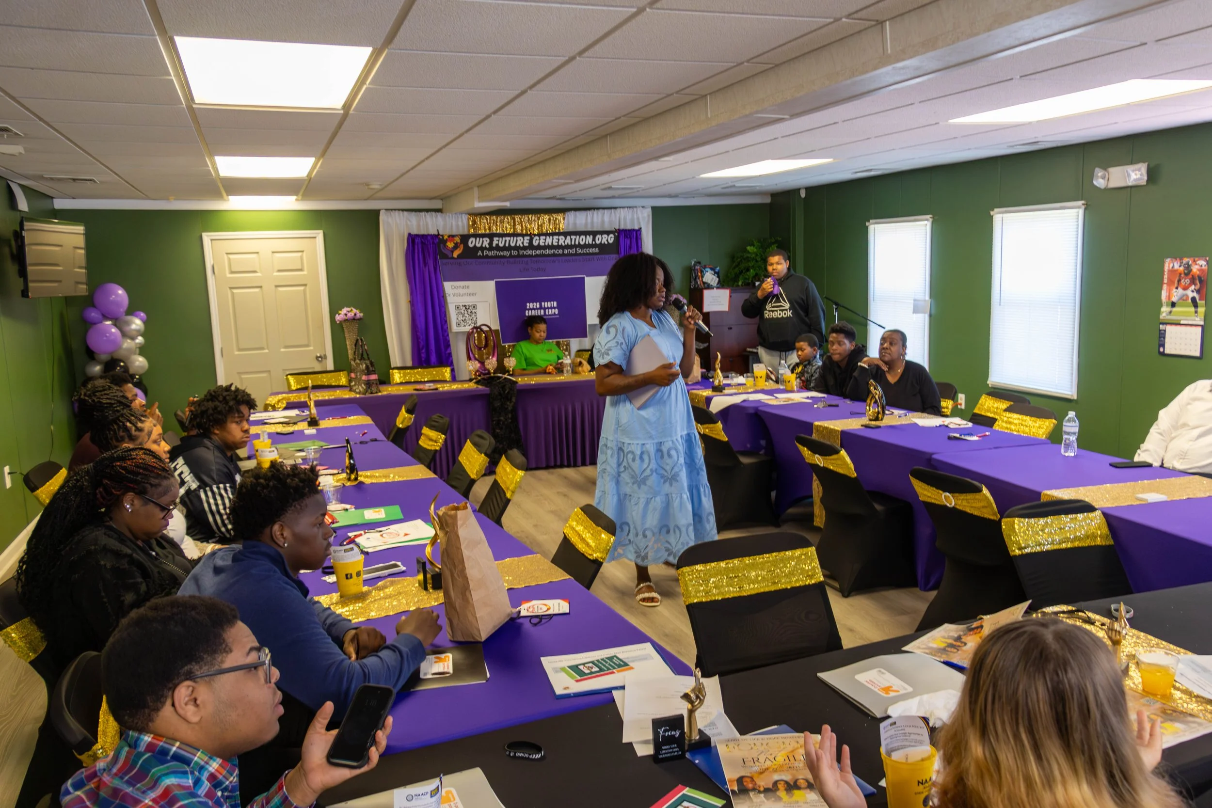 A woman in a light blue dress is speaking into a microphone in a room with green walls and purple tables. Several people are seated around the tables, and a large banner and decorations are visible in the background, indicating a community event or s