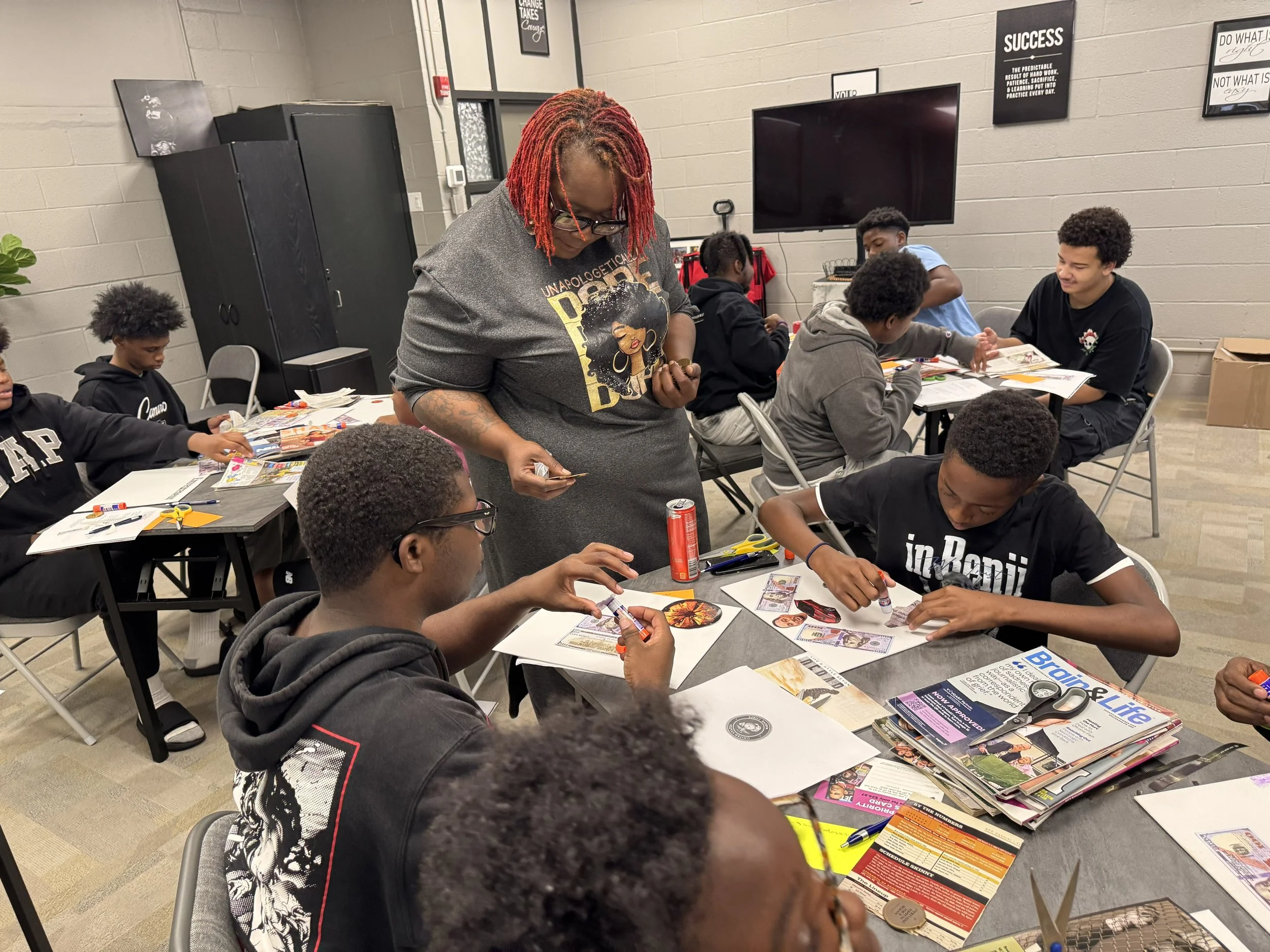 A classroom or workshop setting with multiple students sitting at tables, engaged in a hands-on activity involving colorful paper, scissors, and glue. A woman with red dreadlocks wearing glasses and a graphic t-shirt stands in the center, overseeing 
