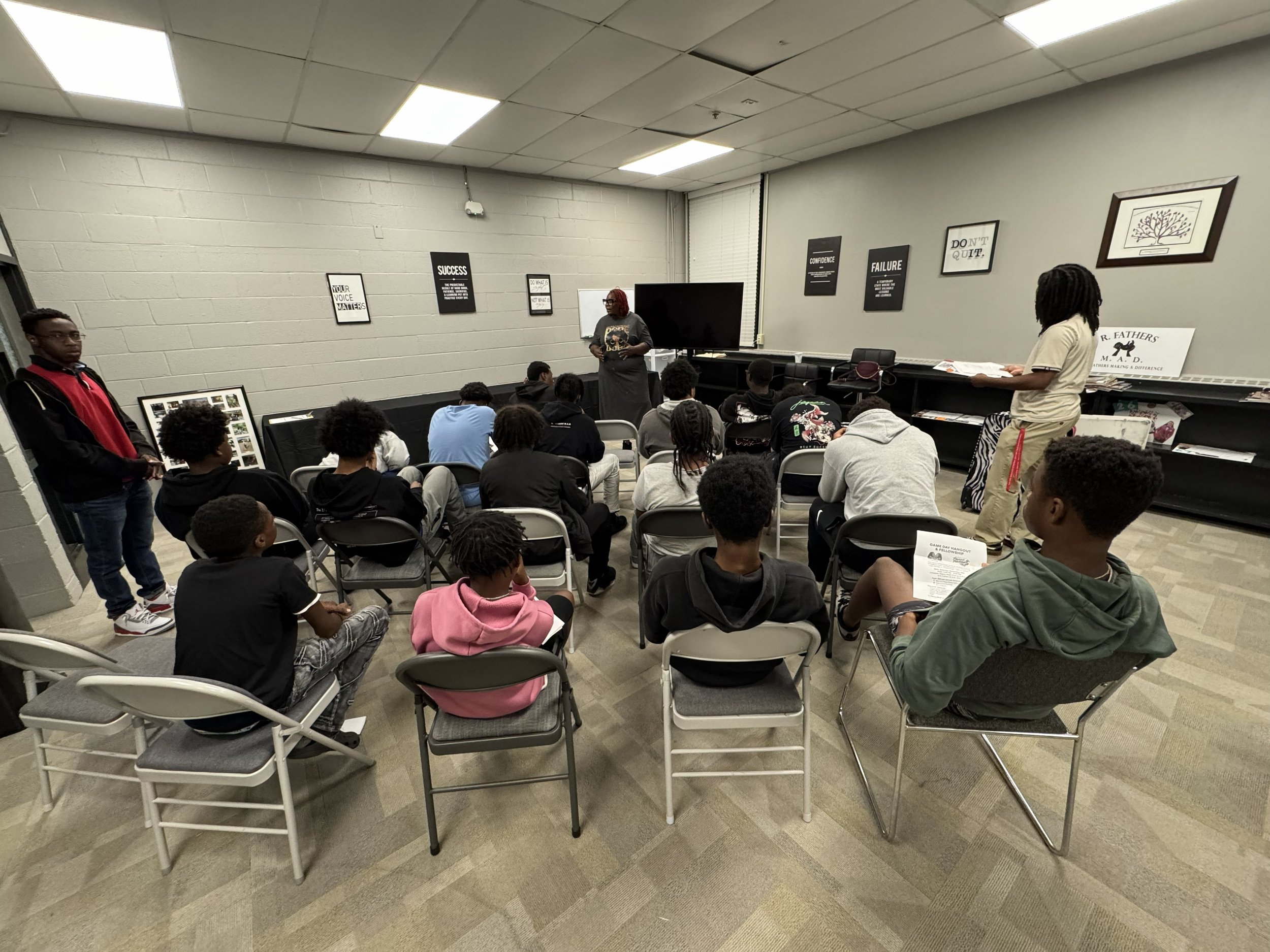 A group of students seated on white chairs listening to a woman standing in front of them in a classroom. The woman is wearing a gray dress with a skull design, and the students are facing her, some taking notes or looking at her. The classroom has p