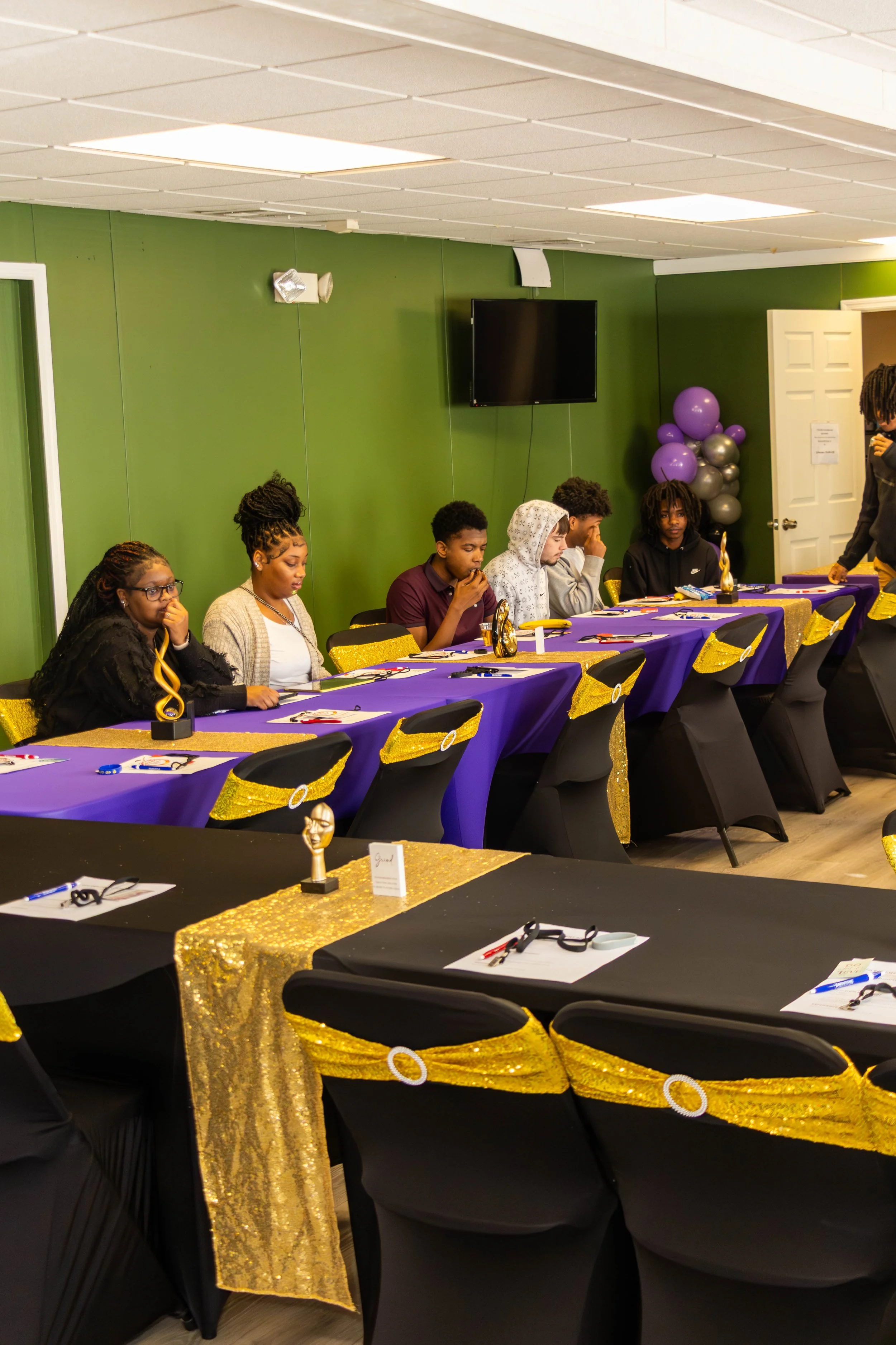 A group of young people sitting at a decorated banquet table. The table is covered with black and purple tablecloths, with gold sequin table runners and gold chair sashes. The people appear to be in a formal setting, possibly a celebration or event, 