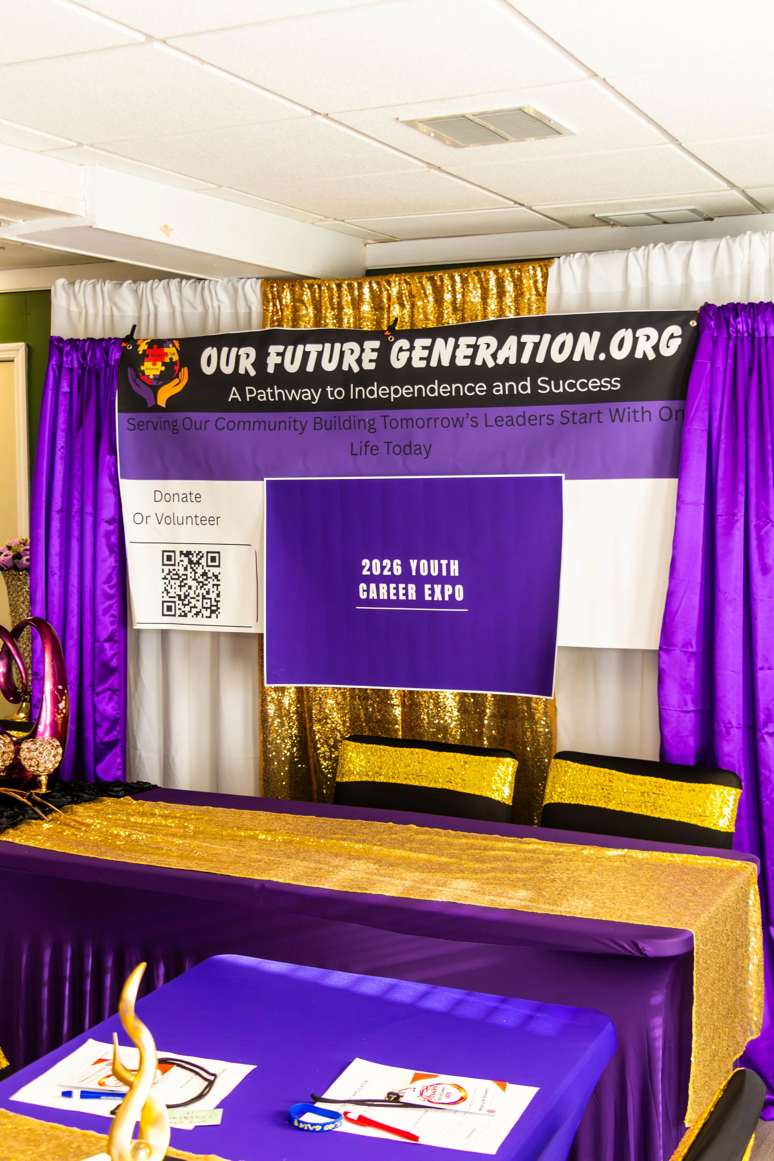 A booth with a black and purple sign that reads 'OUR FUTURE GENERATION.ORG' with a tagline 'A Pathway to Independence and Success.' The booth has purple and white curtains, gold accents, and chairs with black seats and gold backs. There are papers, a