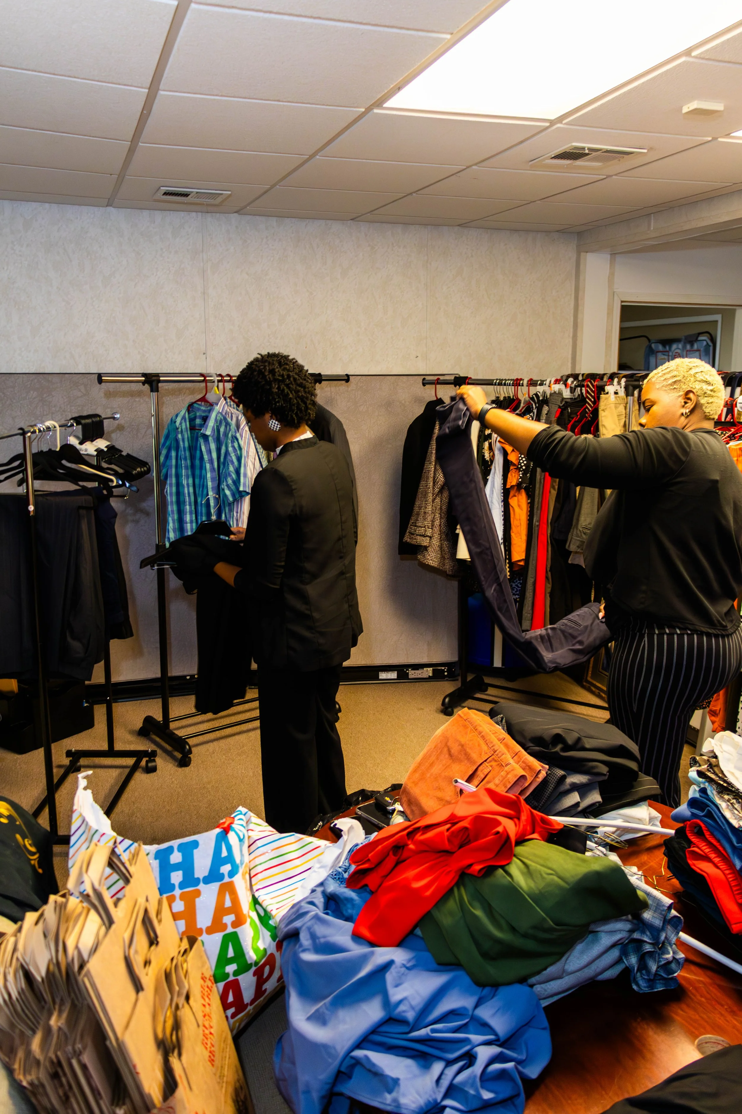 Two women shopping for clothes in a store. One woman with short blond hair is holding a pair of pants, and the other woman with curly black hair is looking at her phone. The store has racks of various clothes and a table overflowing with folded cloth