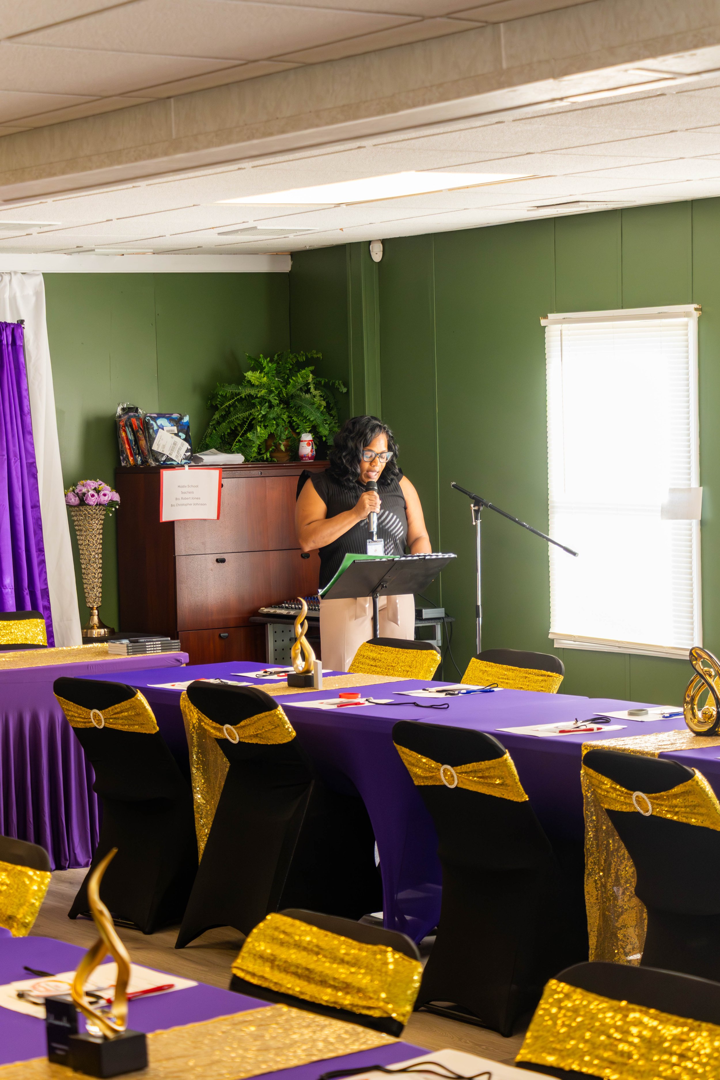 A woman standing at a podium with a microphone in a decorated event room, with tables covered in purple cloth and gold-trimmed black chairs, and a green wall with a window, a large plant, and a cabinet with a sign and some items on top.