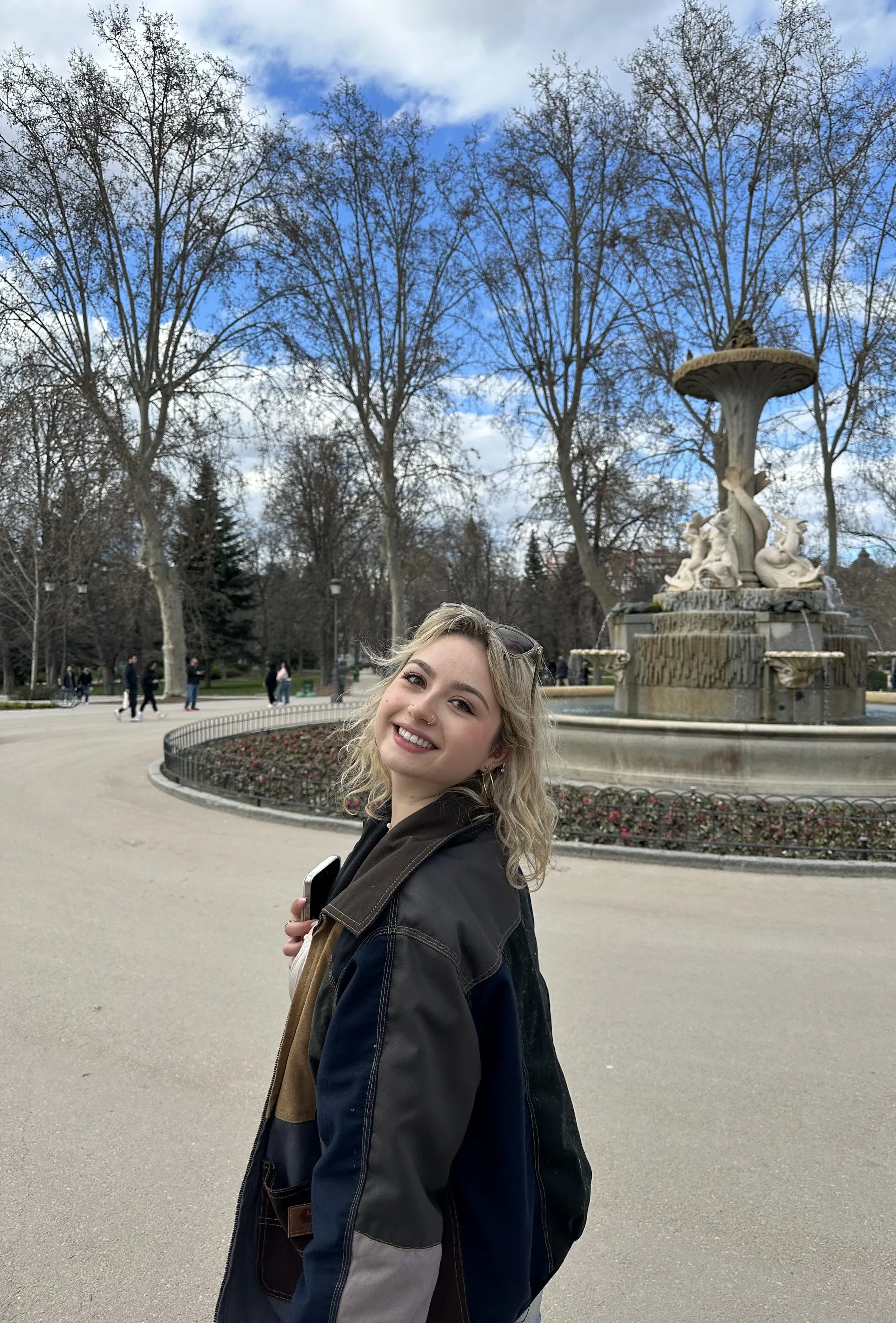 A young woman with blonde, curly hair, smiling and holding a phone, standing in front of a fountain with statues in a park. Trees with no leaves and a partly cloudy sky are visible in the background.