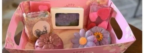 A pink gift box containing various small items including flower-shaped soaps, a small chalkboard sign, and decorative heart-shaped items.